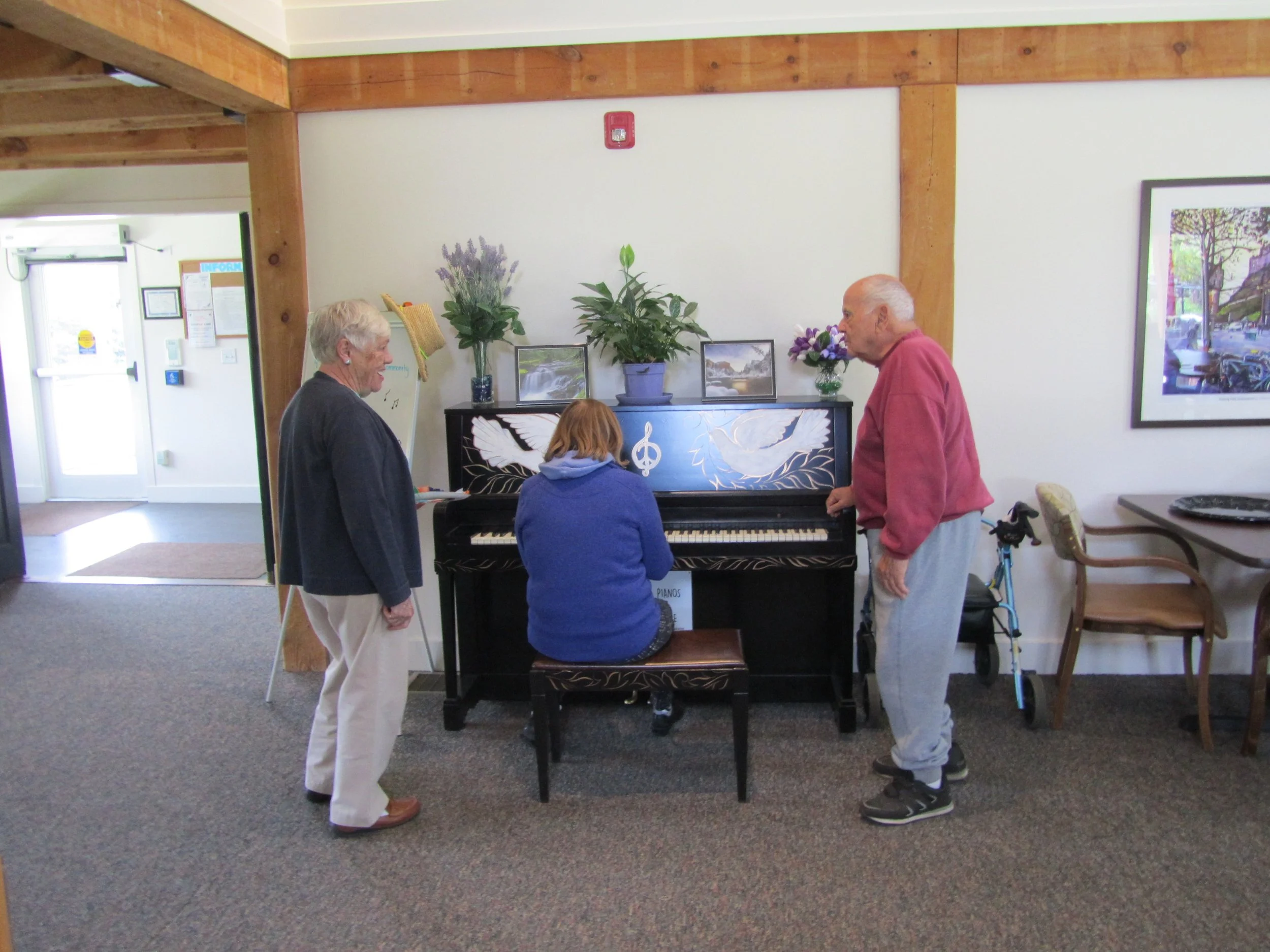Three elderly people gathered around a woman playing a decorated black upright piano, with two framed pictures and flowers on top, in a brightly lit indoor space.