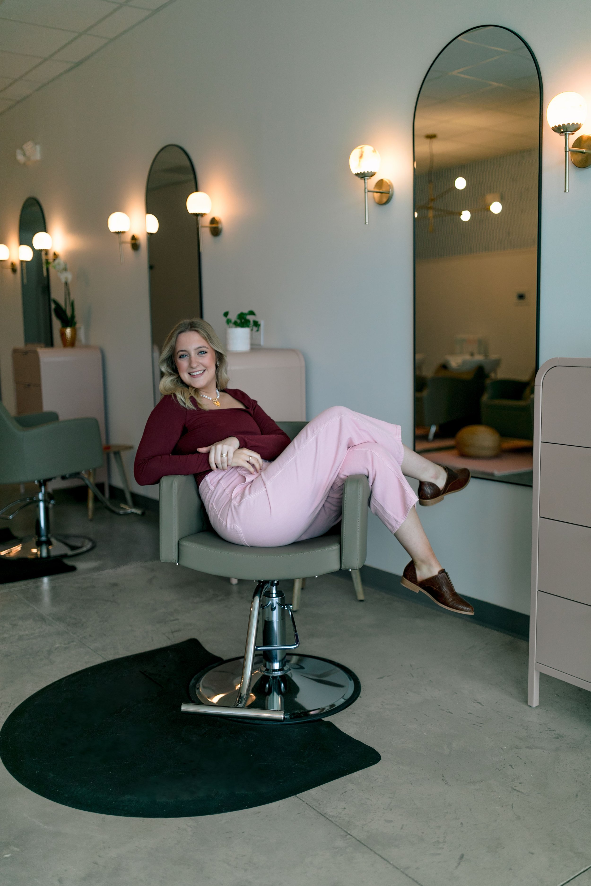 A woman with blonde hair, wearing pink pants and a maroon top, sitting comfortably on a salon chair in a modern hair salon, smiling at the camera.