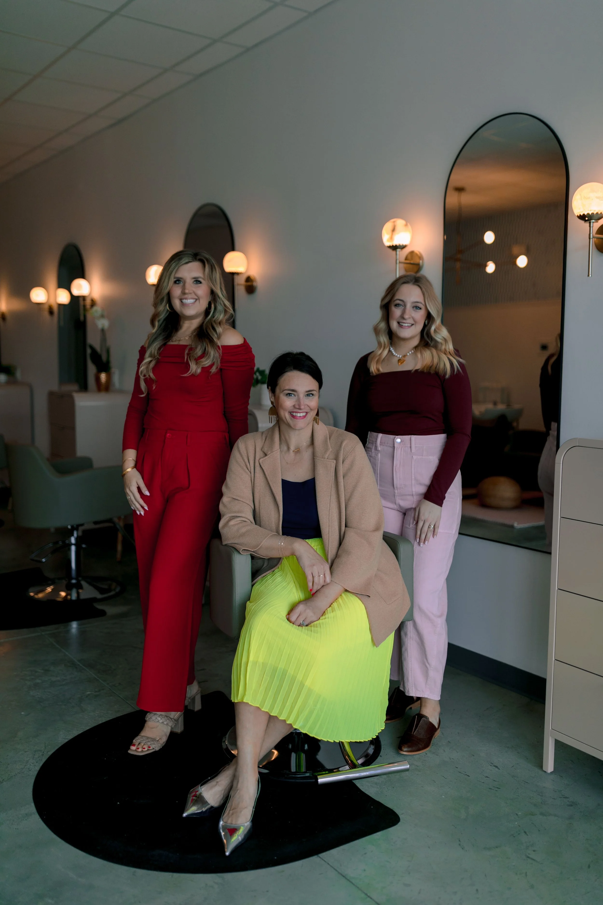 Three women in stylish outfits in a modern salon, one sitting in a styling chair and two standing beside her, smiling.