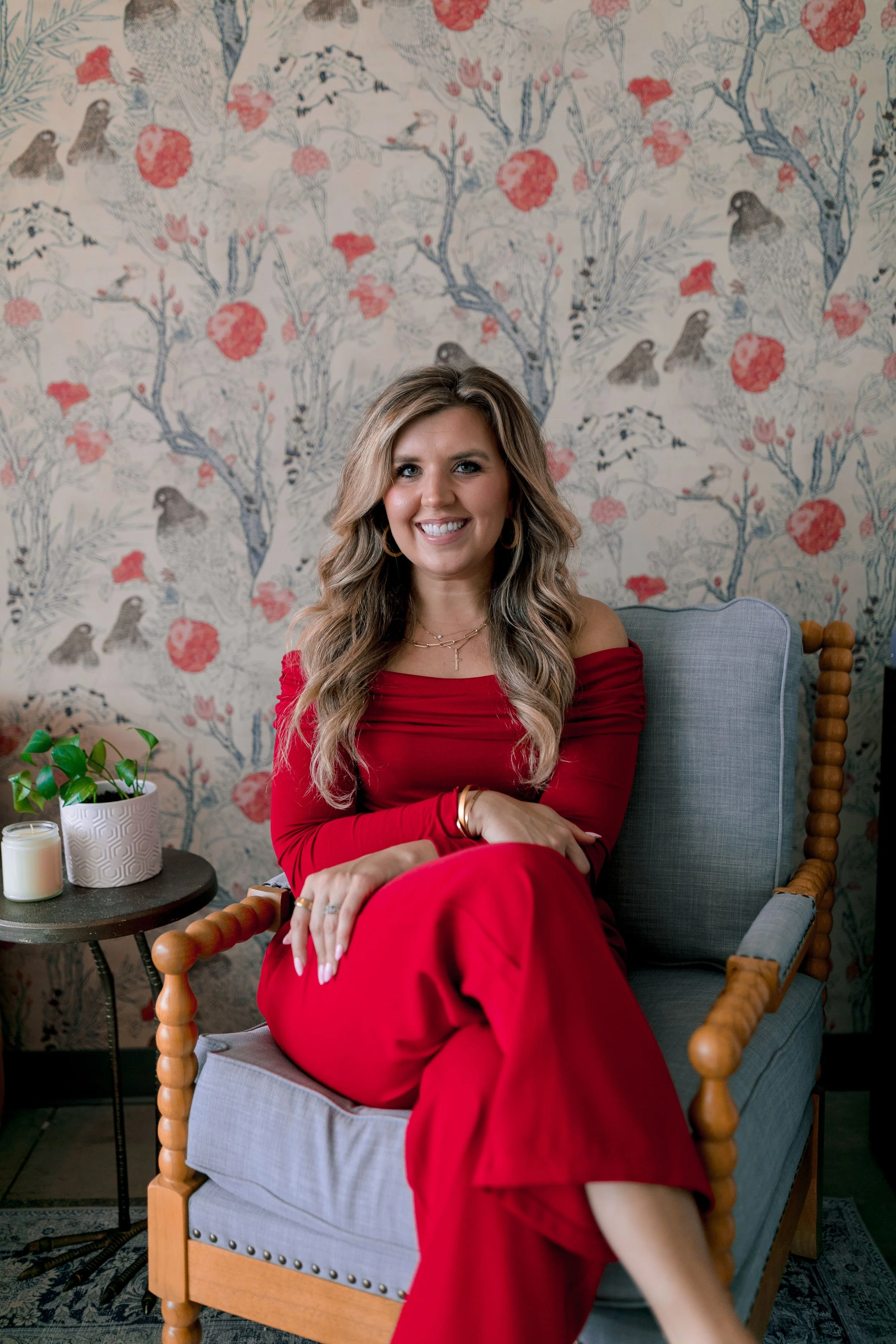 A woman with long, wavy blonde hair sits on a gray upholstered armchair with wooden armrests, wearing a red off-the-shoulder dress, and smiling. Behind her is wallpaper with a floral pattern featuring pink flowers and birds. To her left is a small side table with a potted plant and a candle.