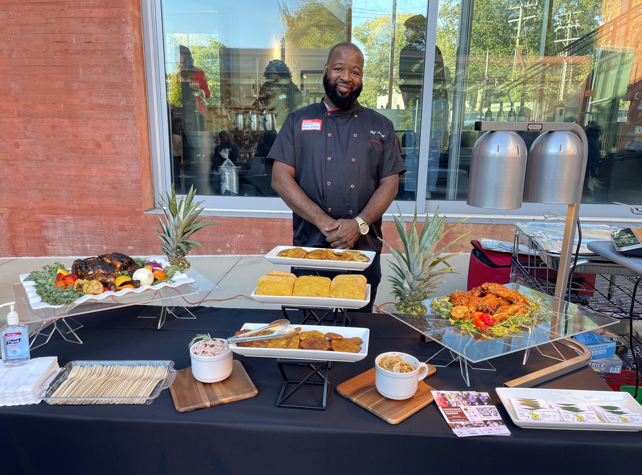 A chef stands behind a table with various plated foods, including roasted chicken, pineapple, and other dishes, set up outdoors in front of a building with large windows.