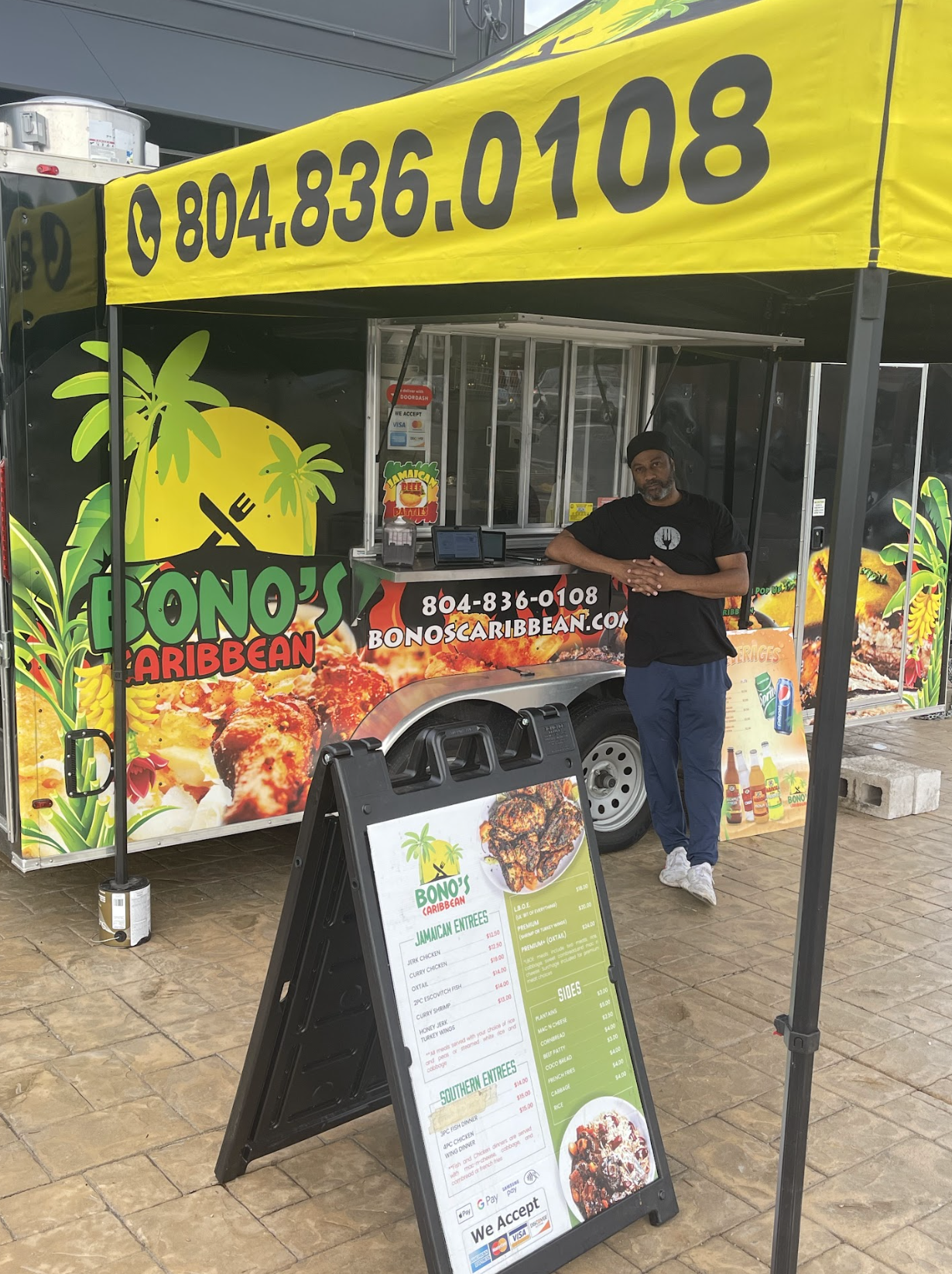 Food truck named Bono's Caribbean with a man standing next to it, a menu board outside, colorful tropical design, and bright yellow canopy with a phone number.