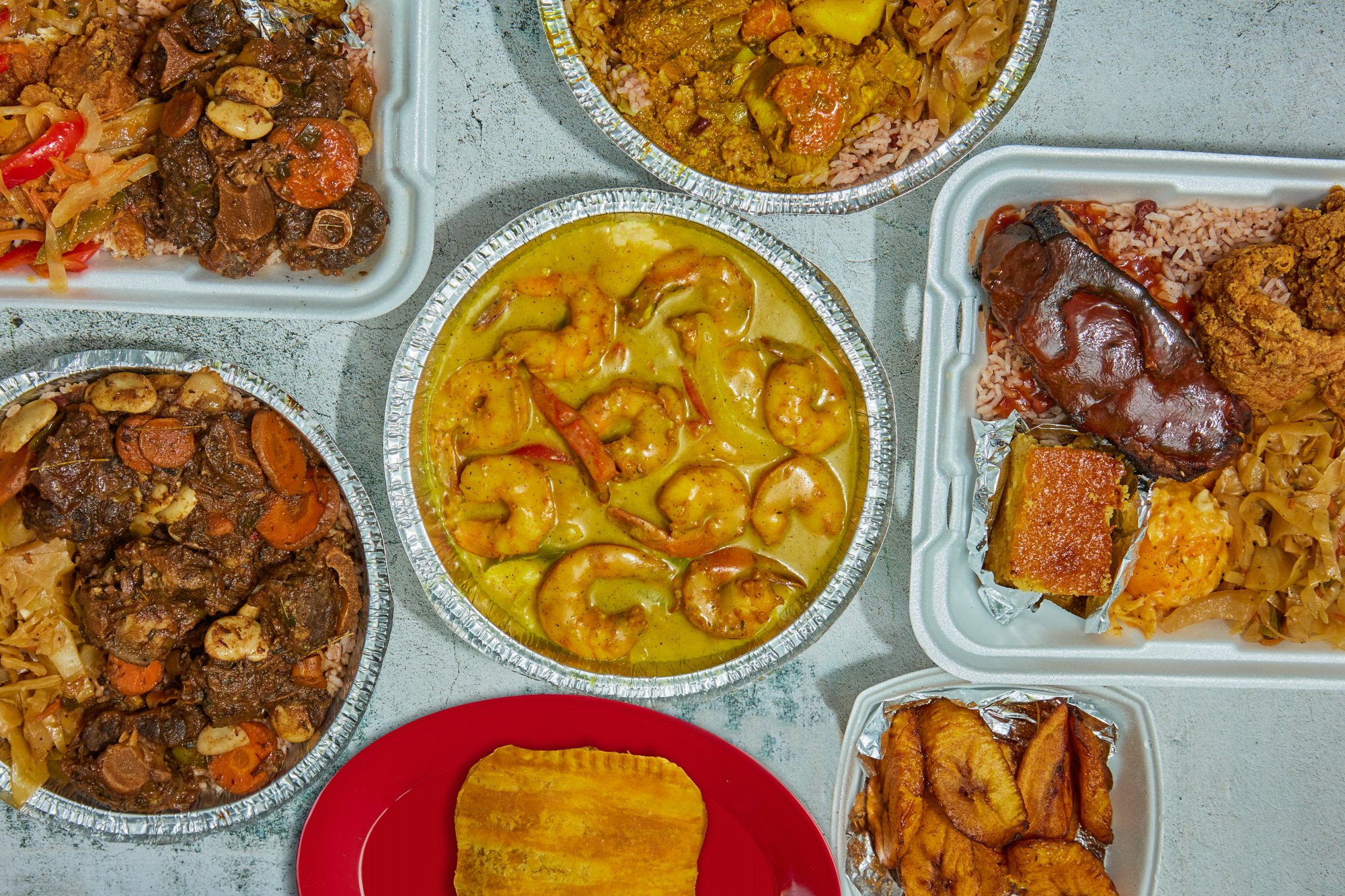 A variety of Indian dishes in foil containers and on a red plate, including seafood curry with shrimp, rice, beef stew with vegetables, fried plantains, and other vegetable dishes.