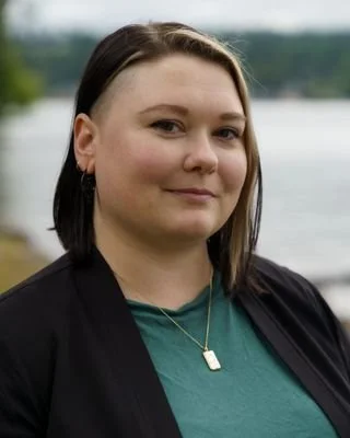 A woman with shoulder-length dark hair, wearing a black jacket and a green top, standing outdoors with a body of water and trees in the background.