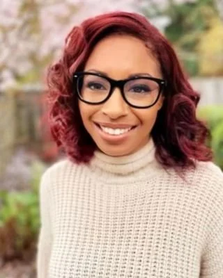A woman with red wavy hair wearing black glasses and a cream-colored turtleneck sweater, smiling outdoors with greenery in the background.