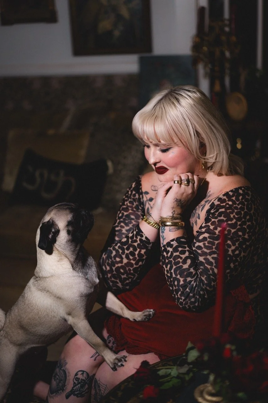 A woman with blonde hair and tattoos, wearing a leopard print top, sits at a table with red candles and holiday decorations. A small pug dog stands on its hind legs, reaching up to her face, while she looks at the dog with her hand resting on her chin.