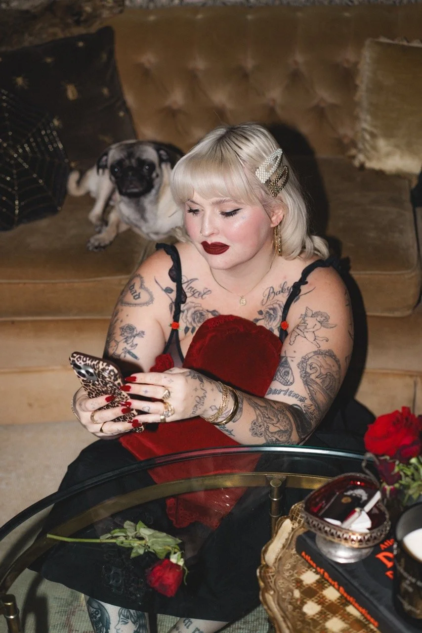 A woman with tattoos and platinum blonde hair, wearing a red velvet dress and red lipstick, sitting on a sofa with a pug dog behind her. She is looking at her phone with a leopard print case. The setting appears to be cozy with decorative cushions, a glass table with roses, and a gold-colored tray with various items.