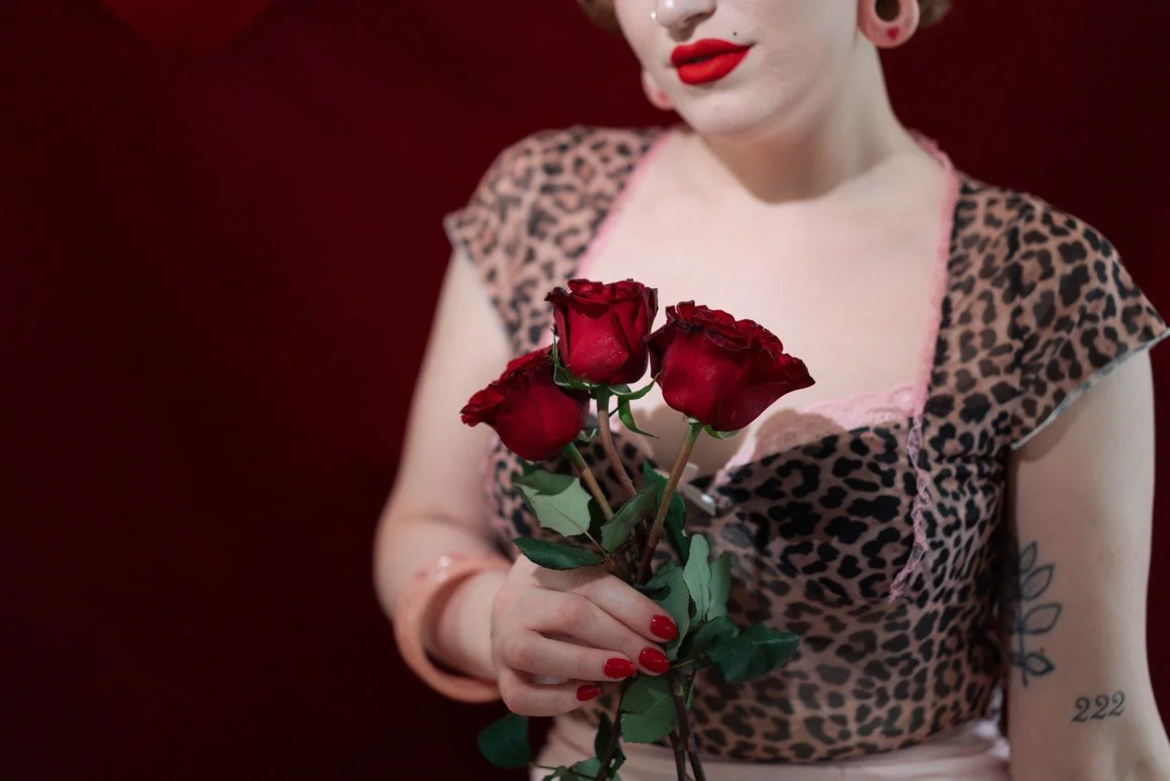 A woman with tattoos and bright red lipstick holding a bouquet of three red roses, wearing a pink leopard print top against a dark background.