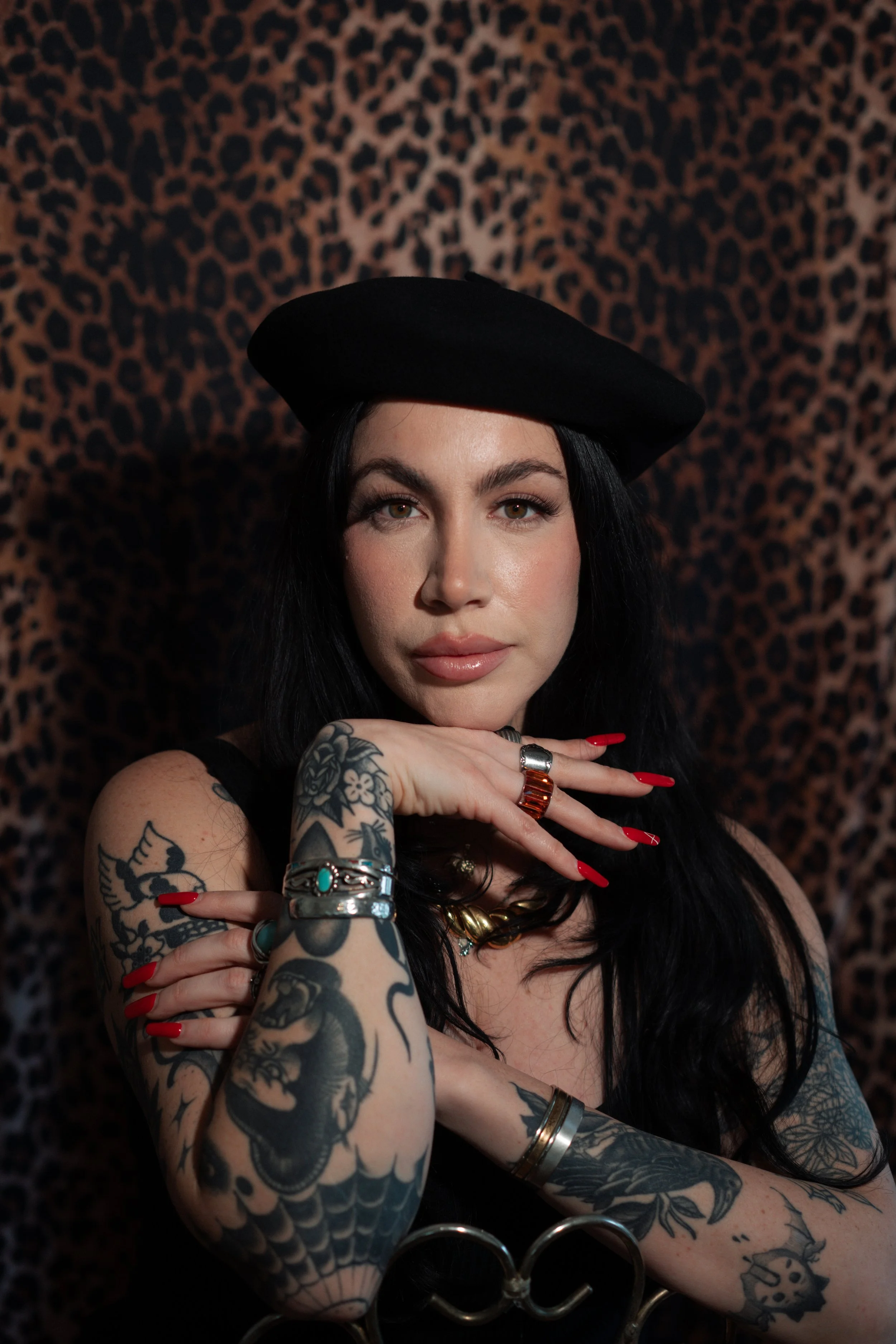 A woman with black hair, tattoos on her arms, and red nail polish, wearing a black hat and jewelry, poses in front of a leopard print background.