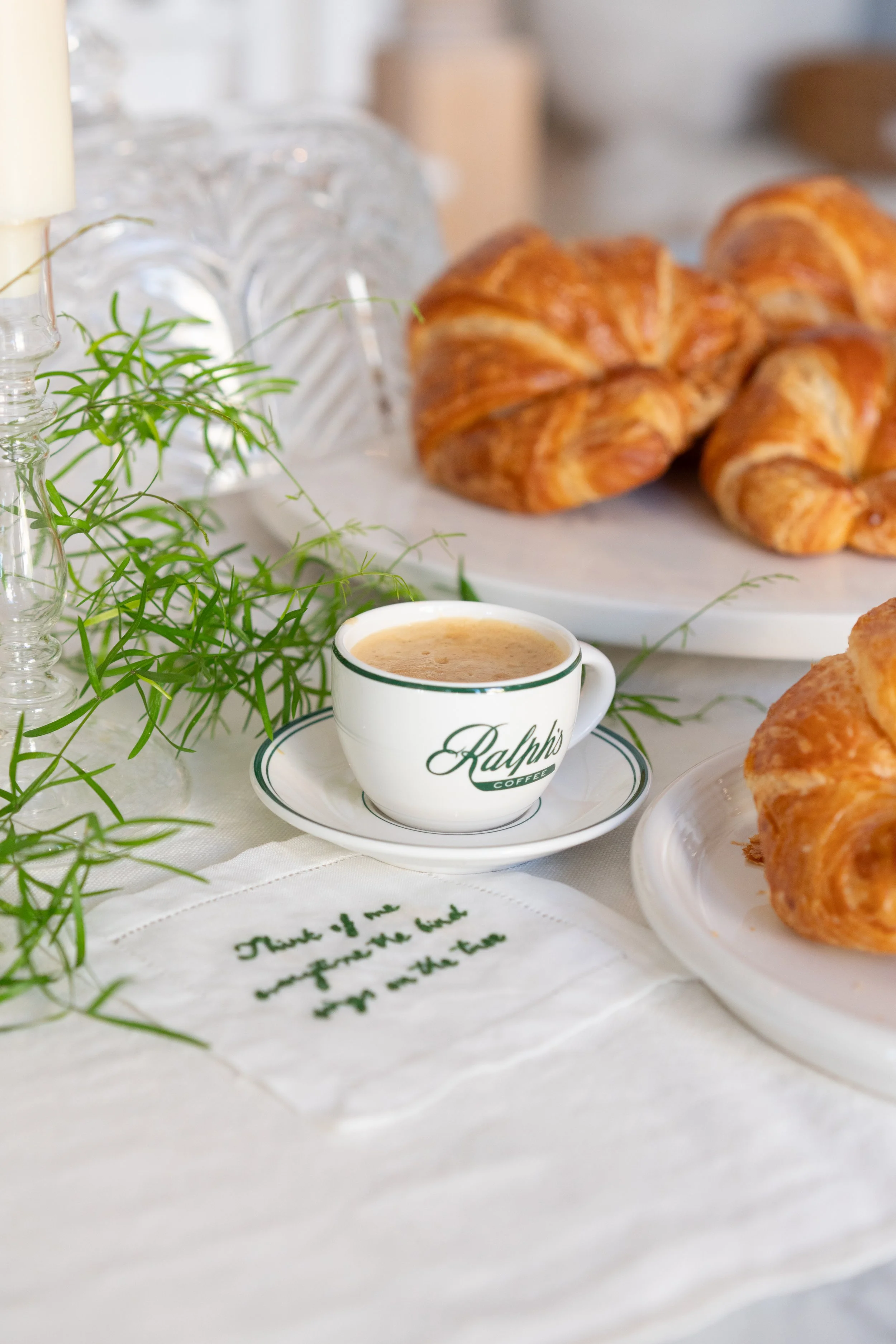 A breakfast scene with a cup of coffee, croissants on a plate, and a decorative plant on a white tablecloth, with a handwritten note.