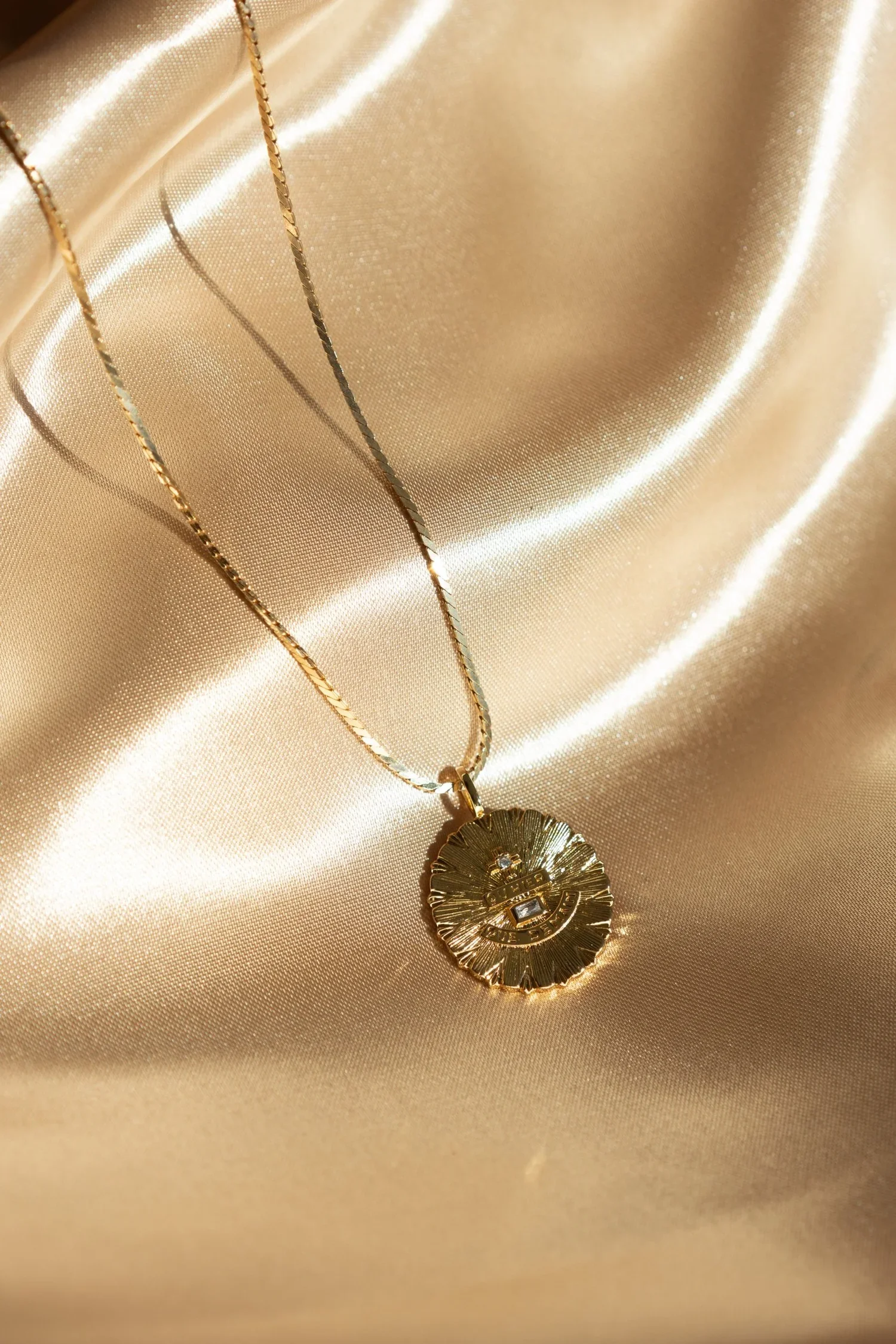 Gold pendant necklace with an engraved circular design resting on shiny, gold-colored satin fabric.