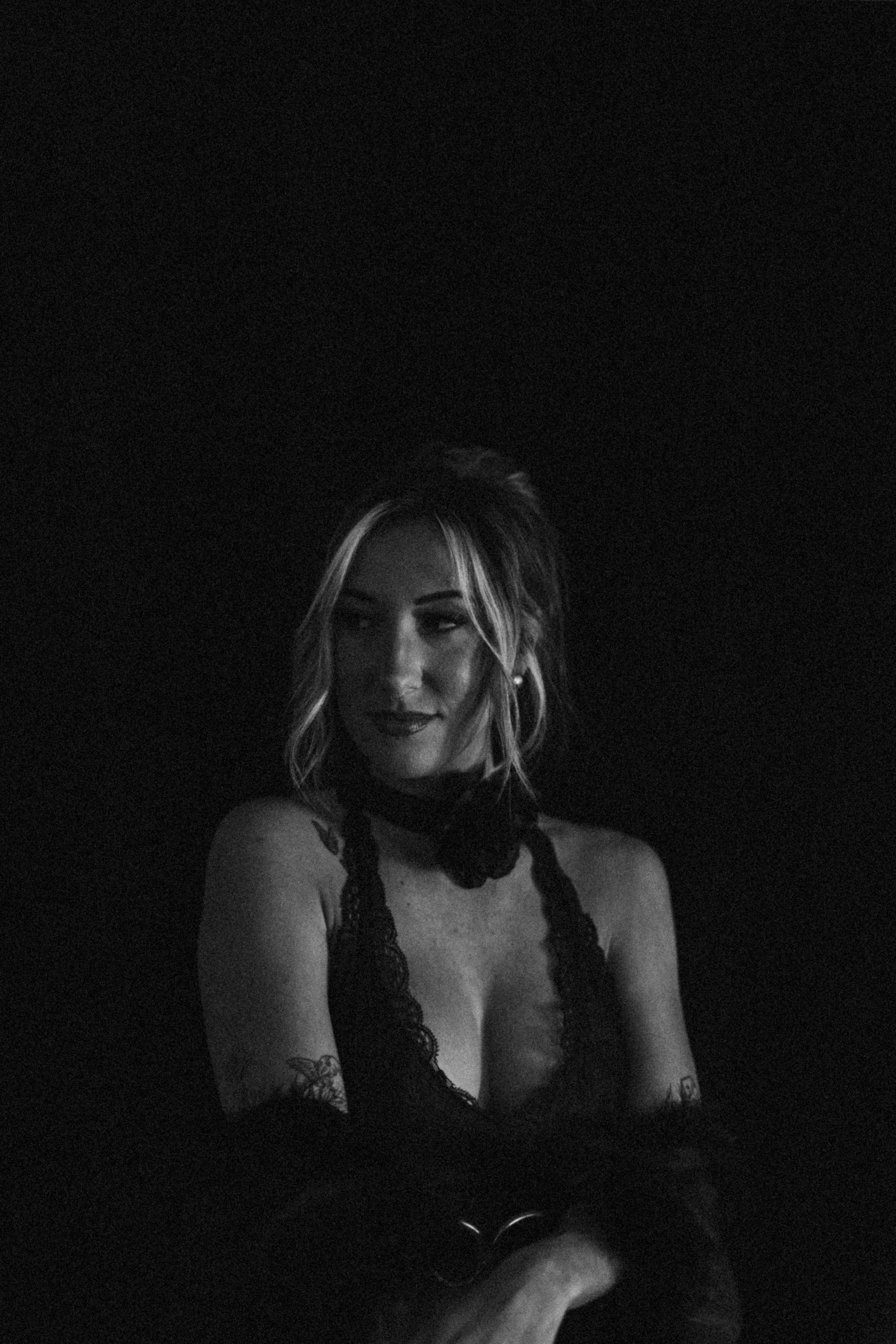 A woman with light-colored hair styled in loose waves, wearing dark makeup and a black lace top, sitting against a black background in a dark setting.