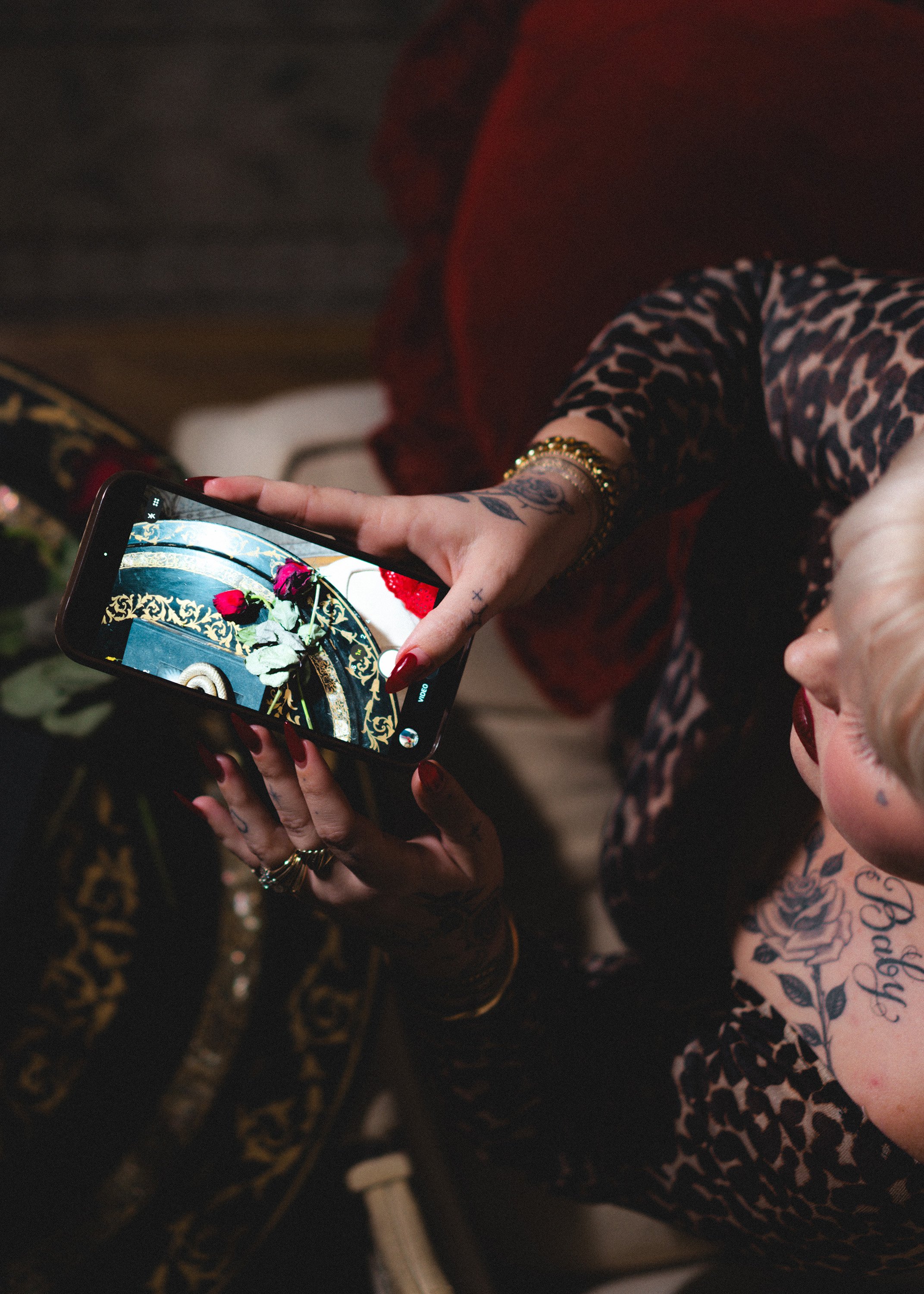 A woman with tattoos and dark red nail polish takes a photograph of a bouquet of red and white roses on a decorative black and gold table with her smartphone. She is wearing leopard print clothing and gold jewelry.