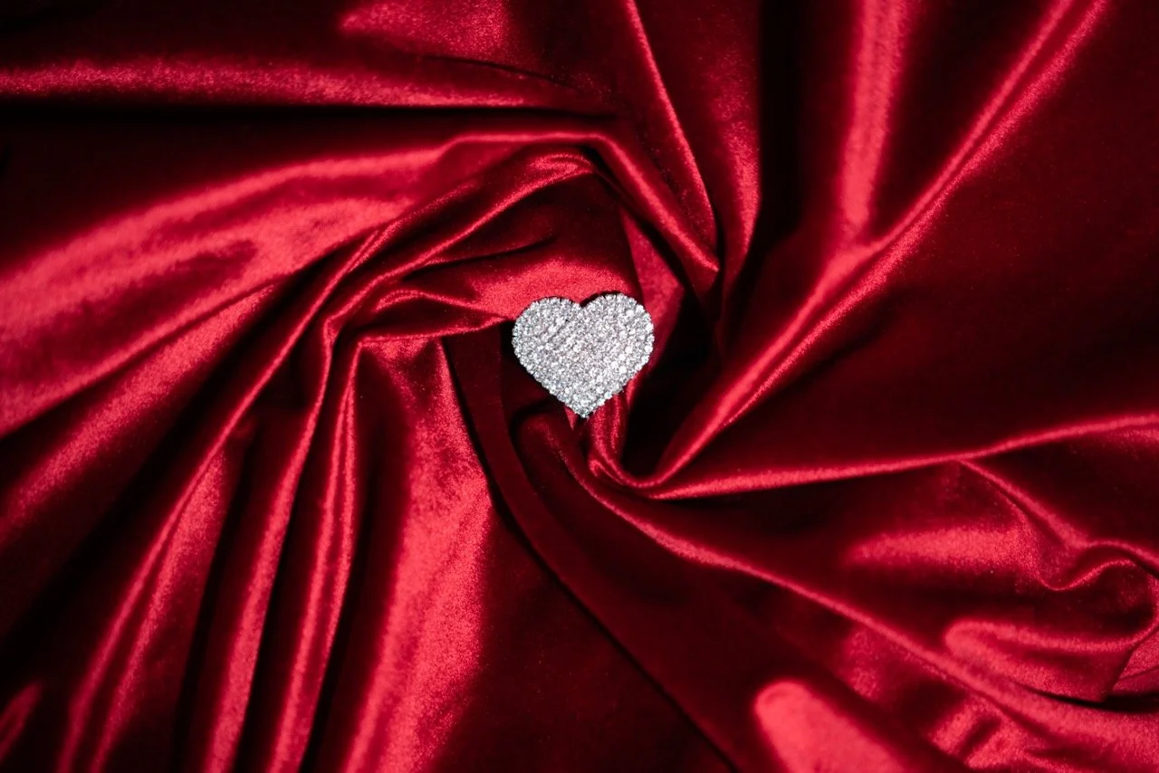 A silver heart-shaped piece of jewelry decorated with small diamonds, placed on red satin fabric.