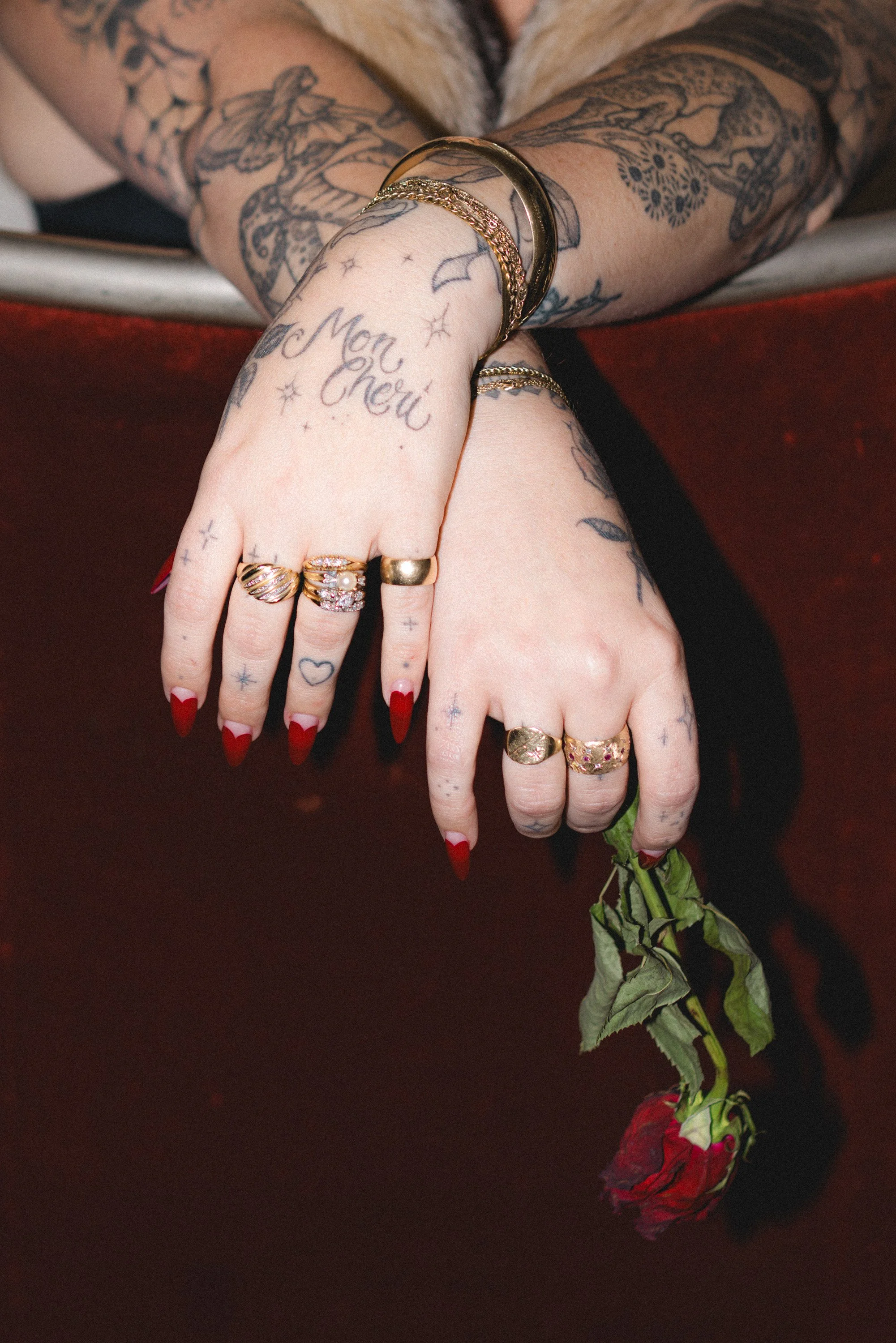 Close-up of a person's tattooed hands with rings and bracelets, holding a wilted red rose against a dark background.