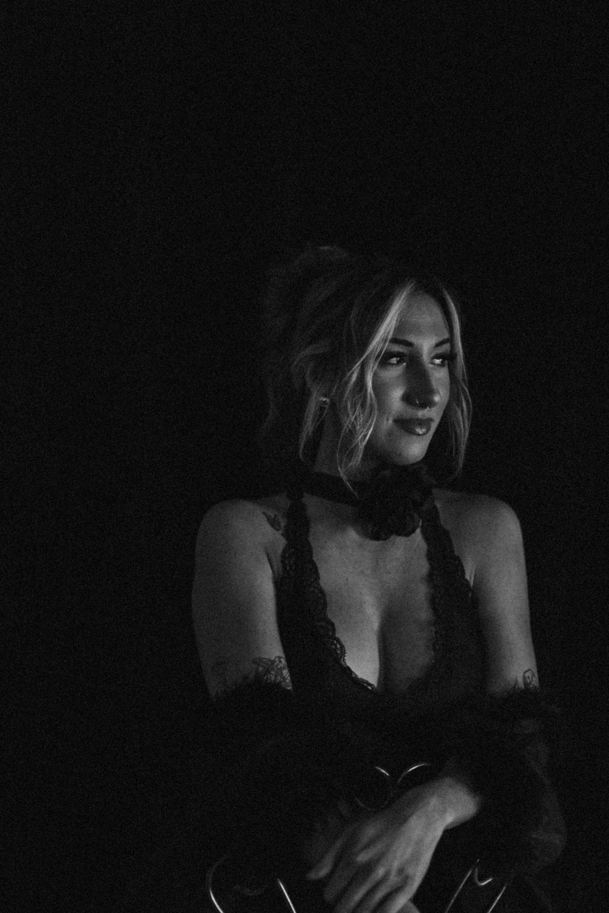 Black and white portrait of a woman with wavy hair, wearing lace clothing, with tattoos on her arms, looking to the side with a slight smile.