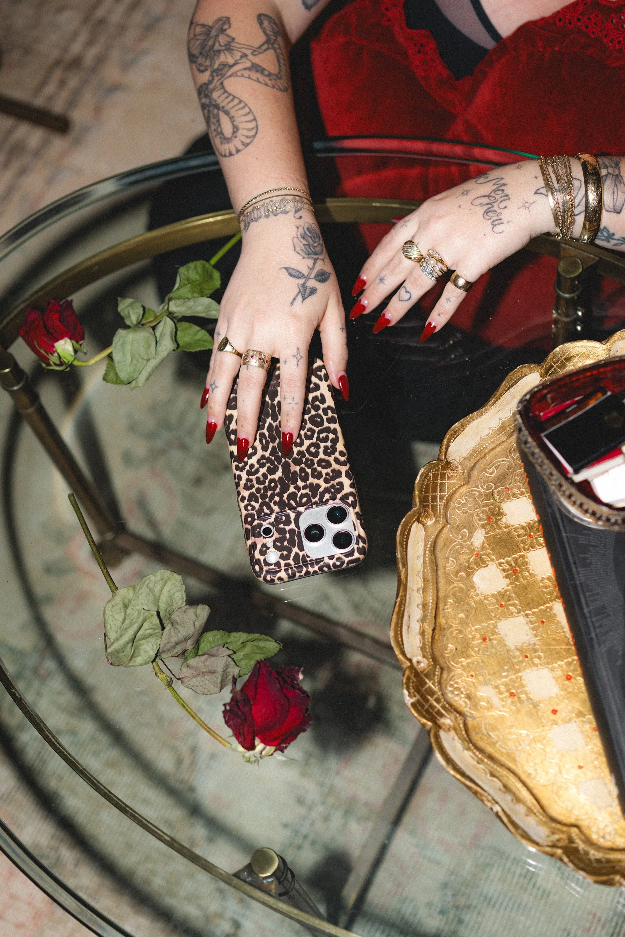 Person with red nail polish and tattoos on their hands, wearing rings and bracelets, holds a phone with a leopard print case over a glass table. On the table, there is a single red rose, a gold decorative tray, and the reflection of a phone.