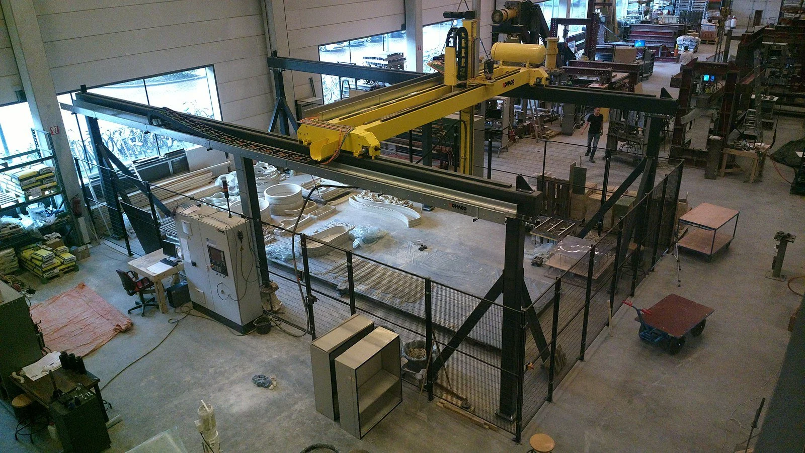View of a large workshop with a crane overhead, surrounding tools, materials, and equipment, and a person walking in the background.