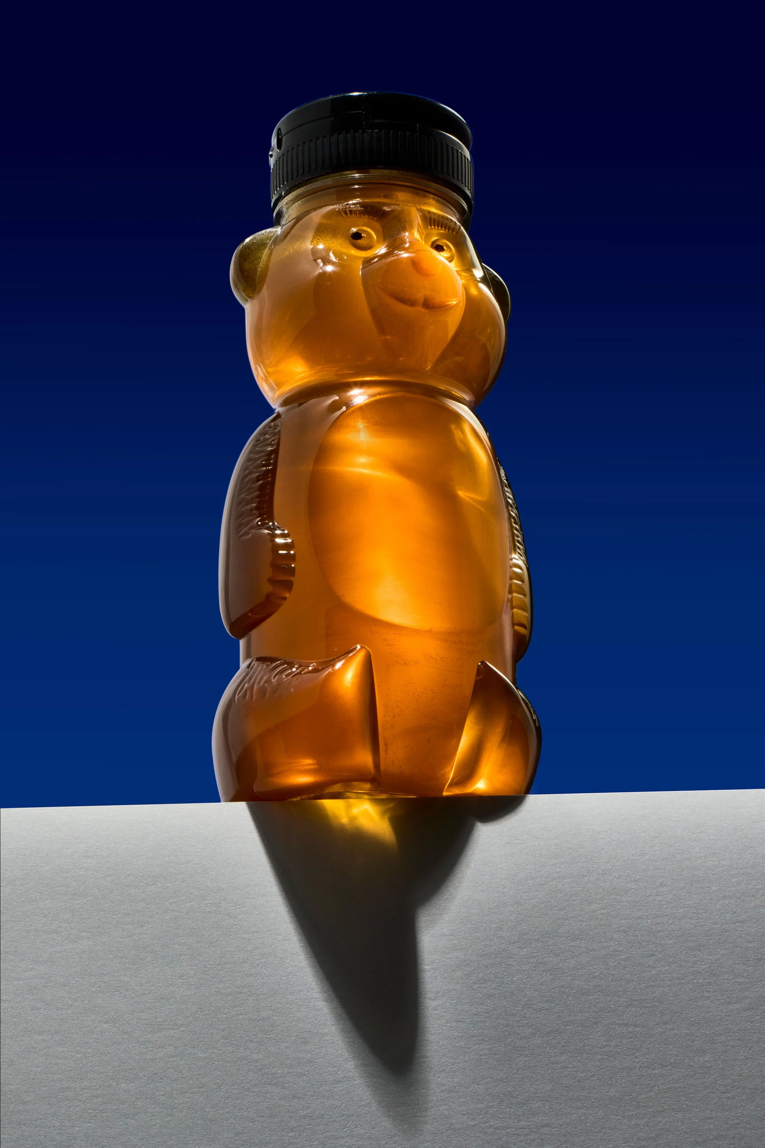 A transparent orange bear-shaped plastic bottle filled with honey, with a black screw-on lid, positioned against a dark blue background and casting a shadow on a white surface.