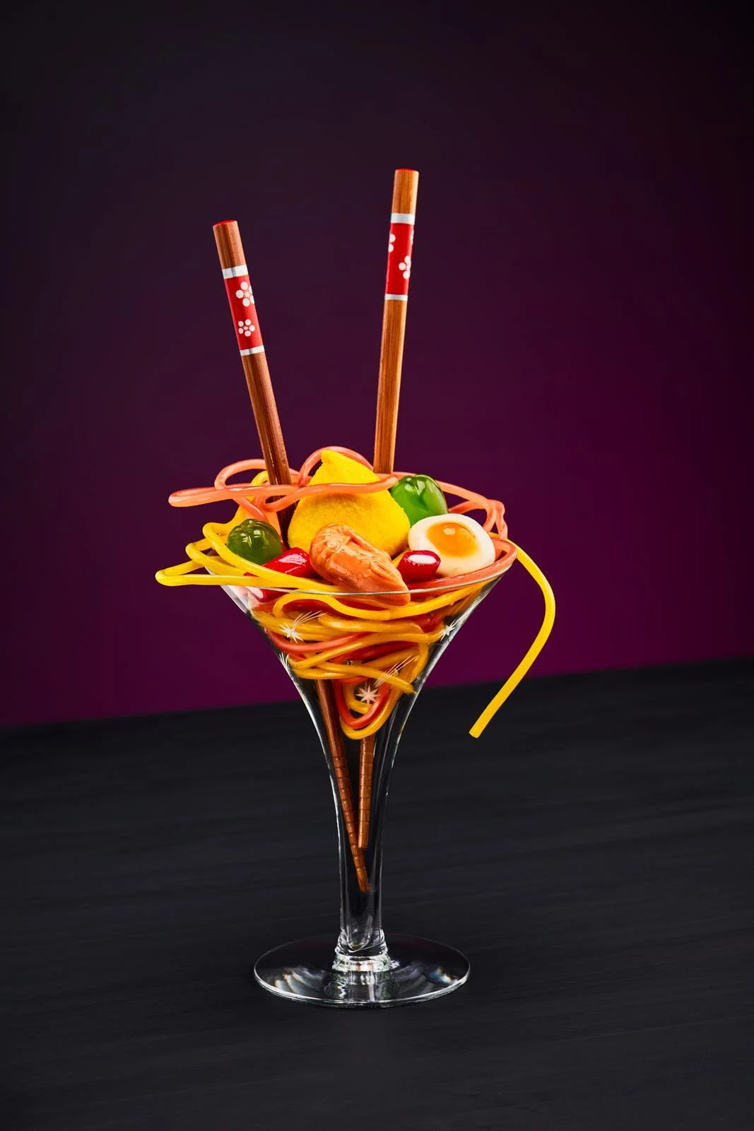 A cocktail glass filled with colorful tangled rubber or plastic noodles, topped with a toy shrimp, a toy boiled egg, cherry, green and red beads, a lemon slice, and two decorative chopsticks.