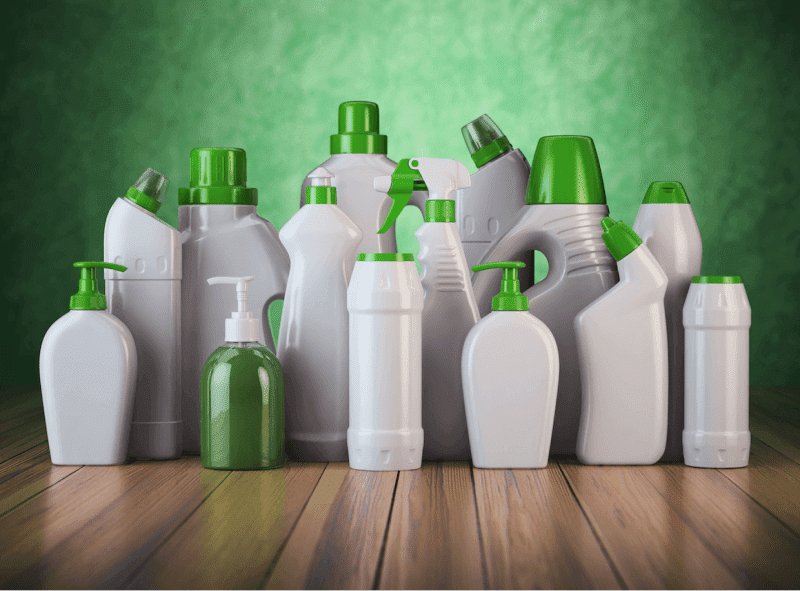 Collection of various cleaning spray bottles and containers with green caps and labels on a wooden surface with a green background.