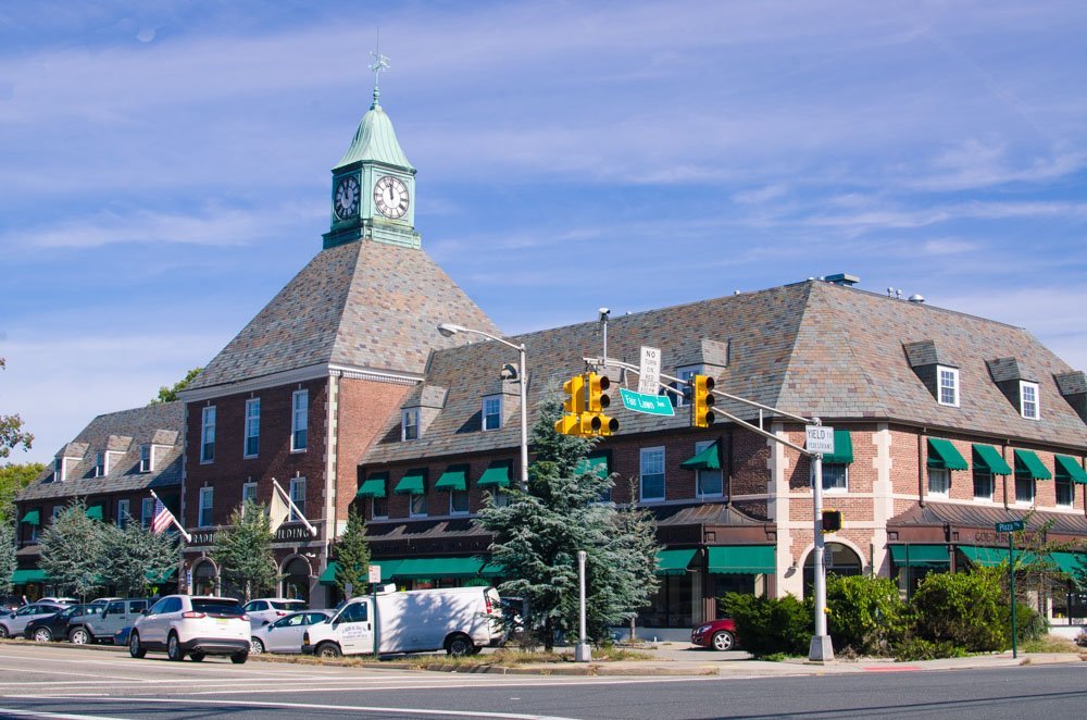 A historic building with a clock tower in Fair Lawn, New Jersey brick facade, green awnings, and surrounded by parked cars and trees at a street intersection. JordyClean offers commercial cleaning service in Fair Lawn, New Jersey.