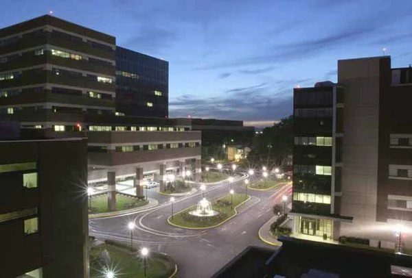 Cityscape at dusk with modern hospital and medical office in Hackensack, New Jersey, a winding driveway, and streetlights illuminating a small central fountain. Janitorial services provided.