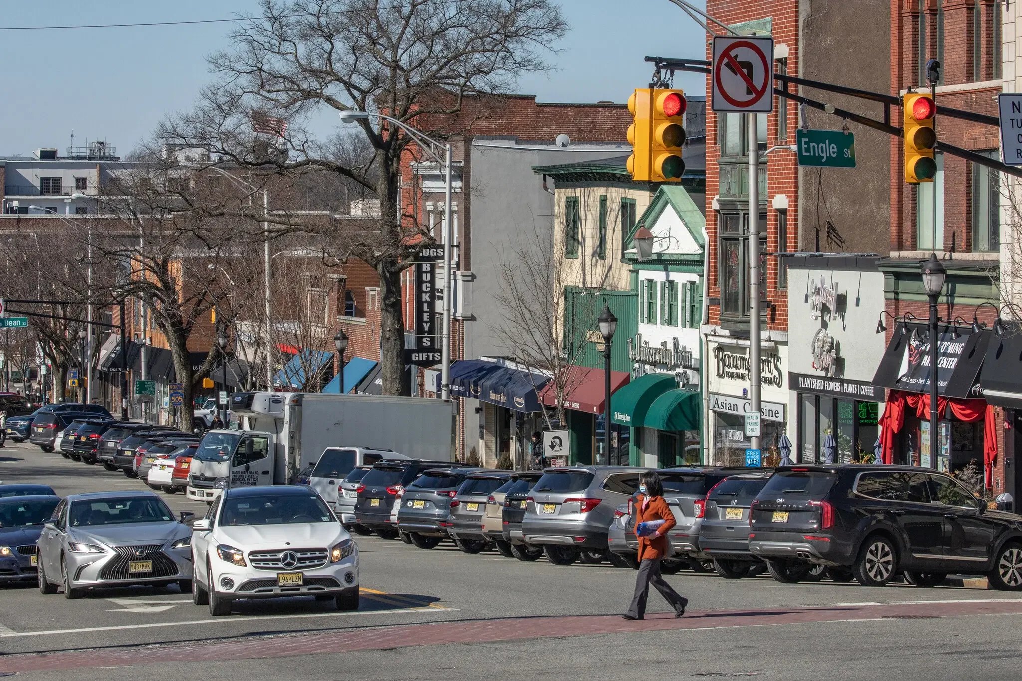 A street scene with parked cars on both sides in Englewood, New Jersey, a woman in a brown coat walking across a crosswalk, storefronts with awnings and signs, leafless trees. JordyClean offers office cleaning service in Englewood, NJ.