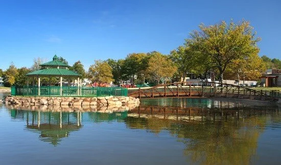 A park scene with a small pond, a gazebo with a green roof, trees, and a wooden bridge over the water on a sunny day in Garfield, New Jersey. JordyClean proudly serves janitorial services to Garfield, New Jersey.
