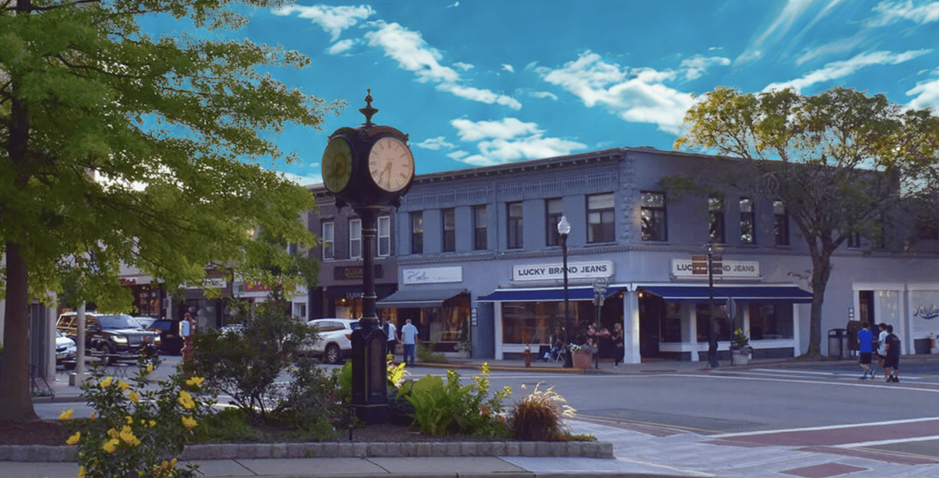 A downtown street scene in Ridgewood, New Jersey, with a clock on a lamppost, trees with green leaves, parked cars, pedestrians, and buildings including a store named 'Lucky Brand Jeans' under a partly cloudy sky. JordyClean provides office cleaning.