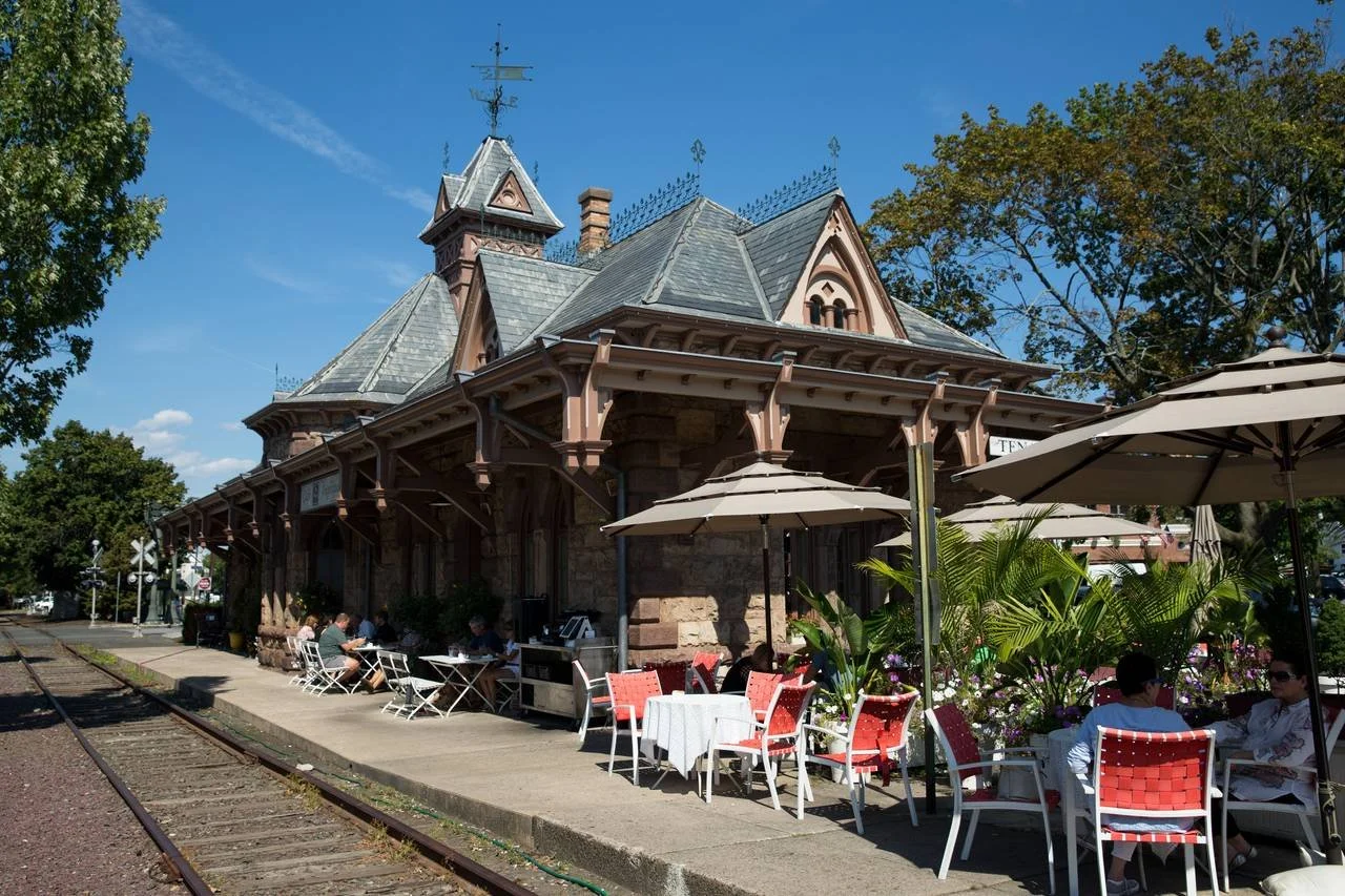Old train station building next to railroad tracks with outdoor seating, tables, and umbrellas on a sunny day in Tenafly, New Jersey. JordyClean offers full commercial cleaning services to Tenafly, New Jersey.