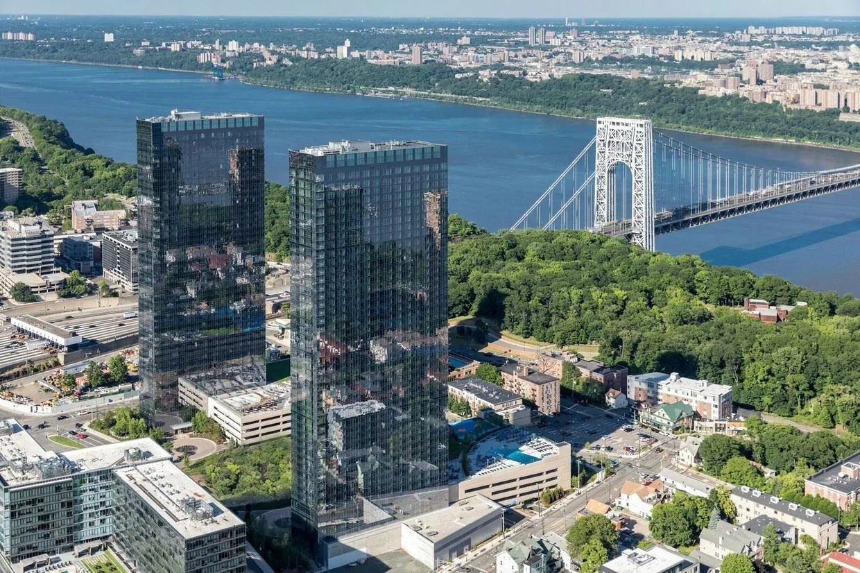 Aerial view of a cityscape in Fort Lee, New Jersey. featuring two tall, glass office buildings, a bridge crossing a river, and greenery along the riverbanks with a city skyline in the background. JordyClean provides commercial cleaning in Fort Lee NJ