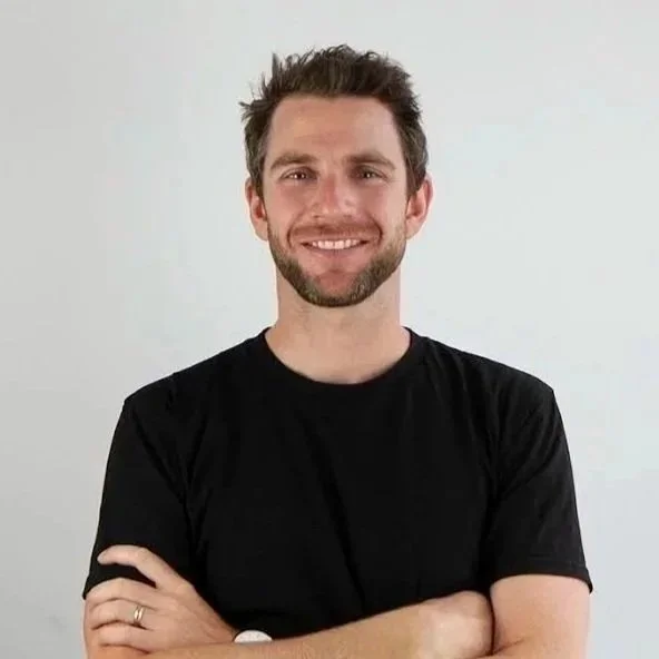 A man with short brown hair and a beard, smiling, wearing a black t-shirt, crossing his arms, standing against a plain white background.