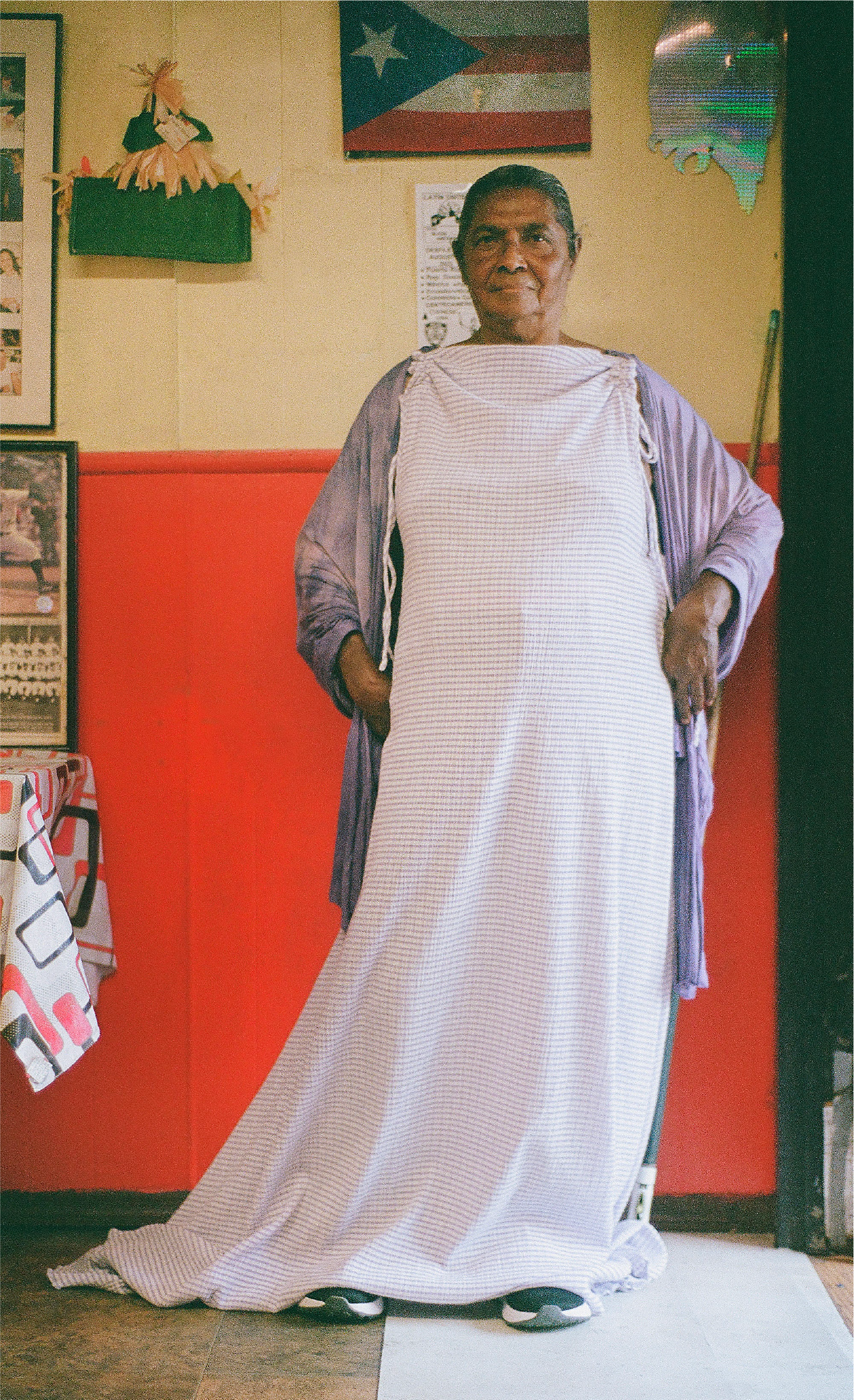 An older woman is standing indoors, wearing a purple gingham, floor-length nightgown. She is wearing a purple shawl and has her hands in her pockets, standing in front of a wall decorated with a Puerto Rican flag and other items.