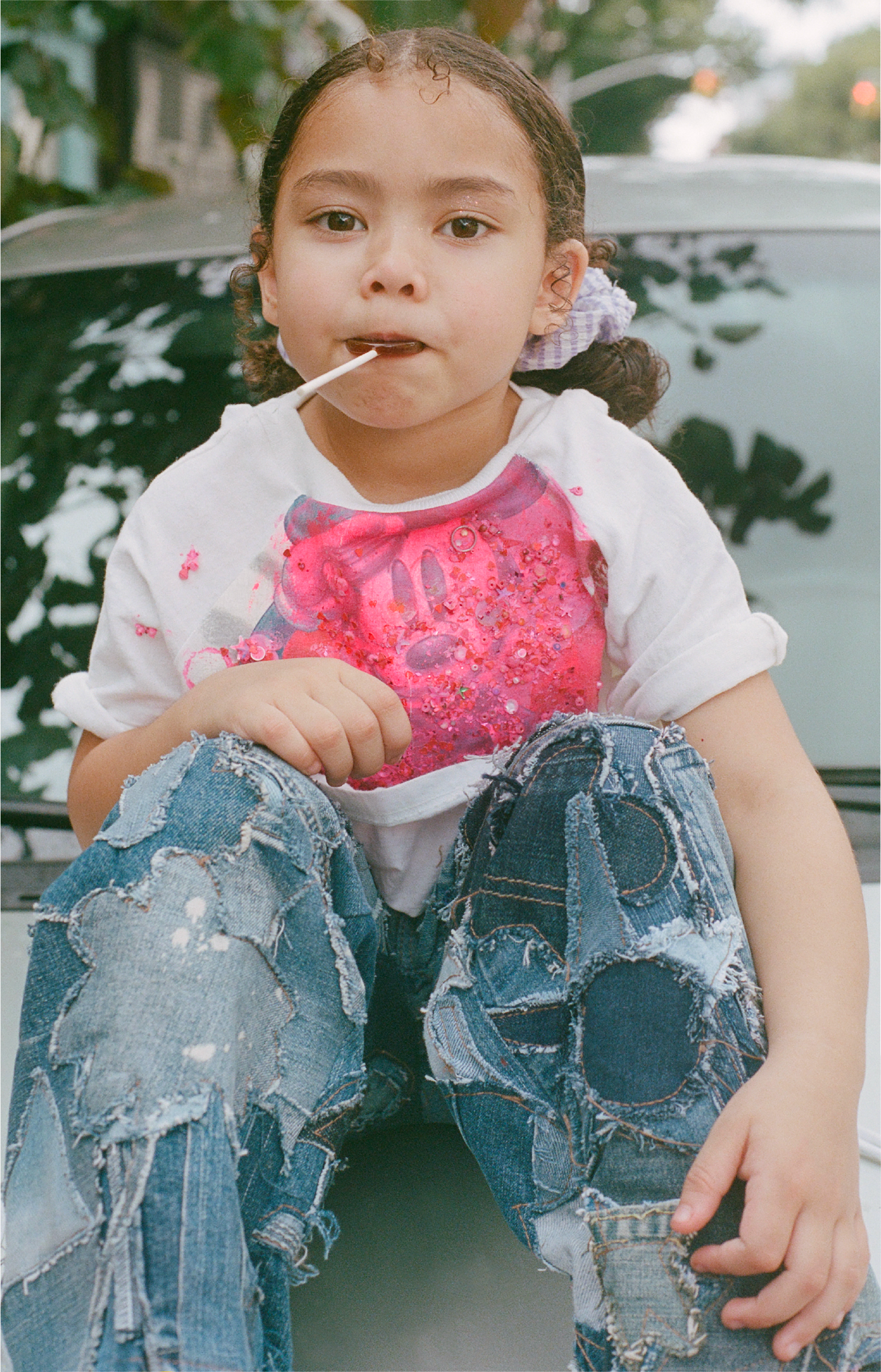 A young girl with curly hair and a purple hair bow sitting on a car, wearing a white T-shirt with a pink graphic and patchwork jeans, holding a lollipop and looking at the camera.