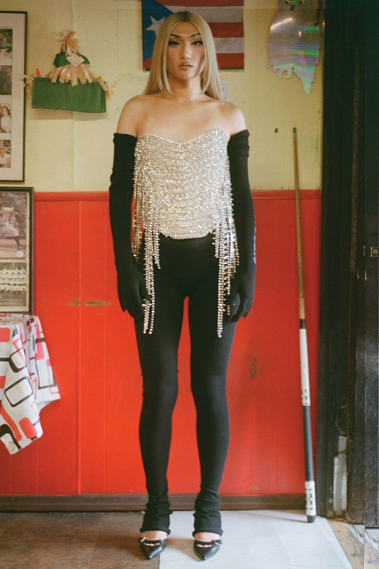 A woman standing indoors wearing a glittering top adorned with rhinestones or crystals, black long gloves, black pants, and high heels, with a flag of Puerto Rico hanging on the wall behind her.