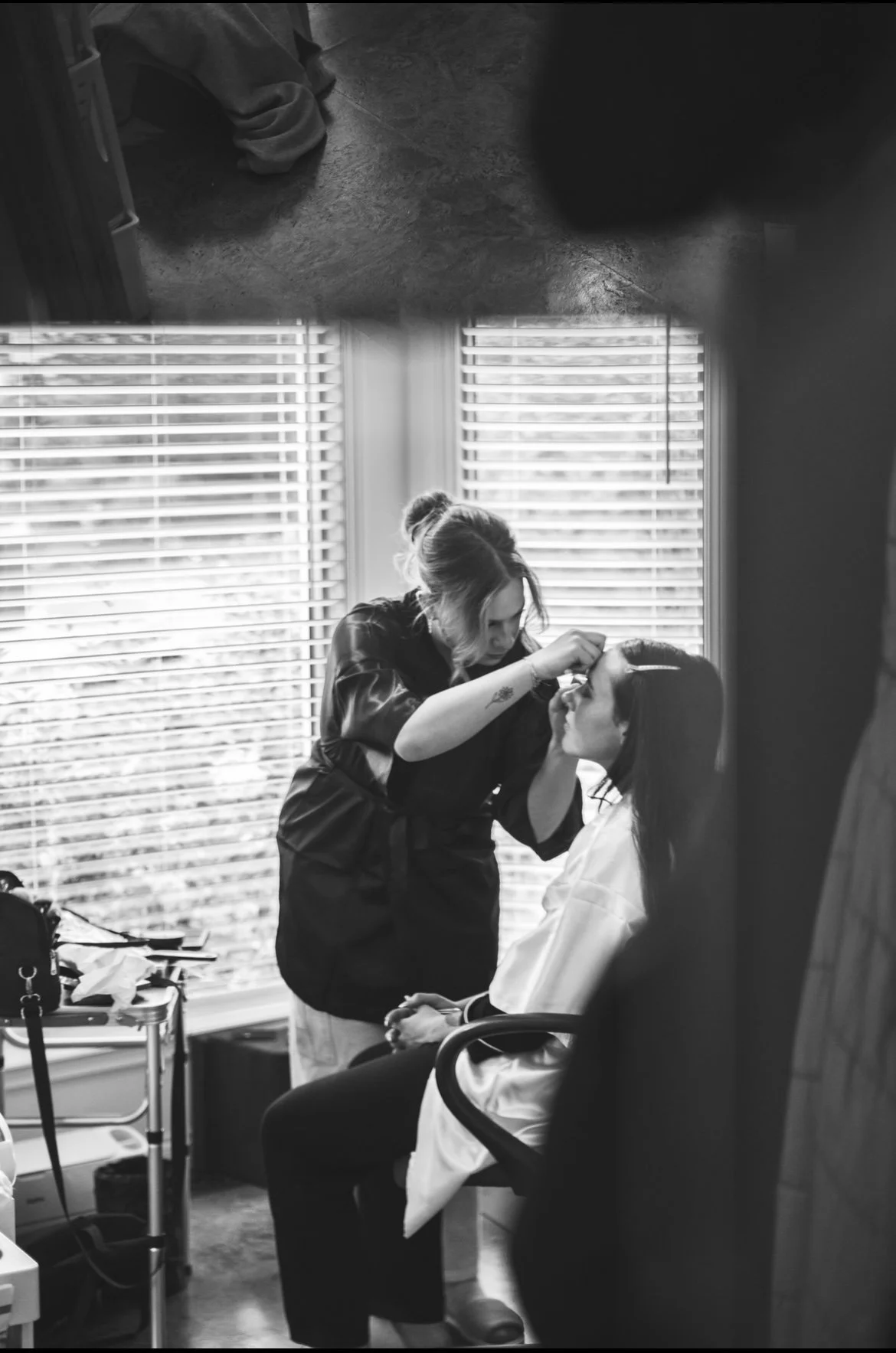A woman sitting on a chair with a makeup cape is having her makeup done by another woman in a room with blinds on the window.