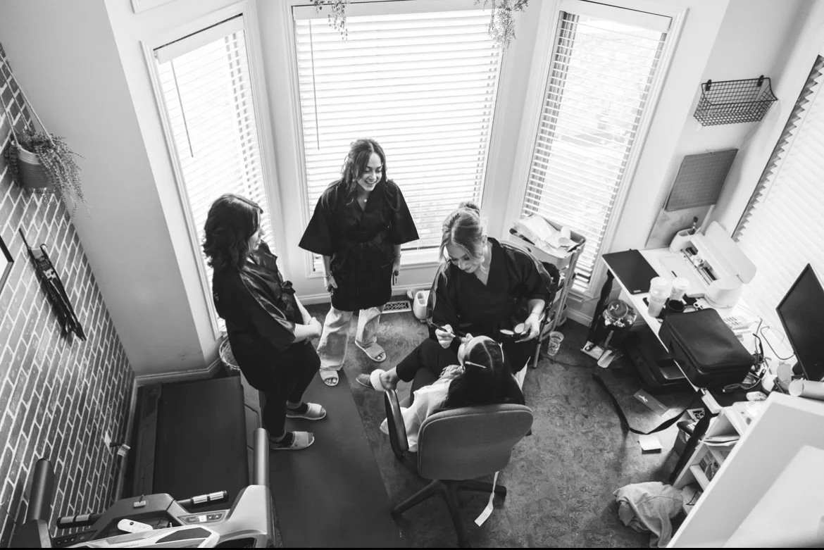 A woman sitting in a chair with three women standing around her in a small room with large windows, a desk with medical equipment, and a treadmill.