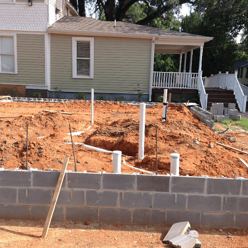 A construction site in front of a house with an ongoing foundation or plumbing installation, showing exposed pipes and a low concrete block wall.