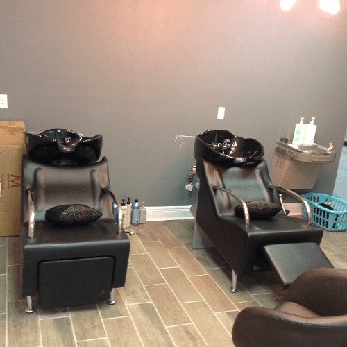 Two black salon shampoo chairs with sinks in a hair salon, a wall with outlets, and a blue laundry basket in the background.