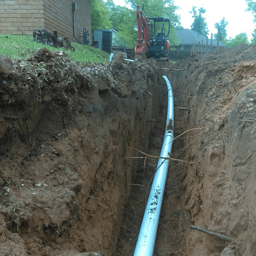 A construction site showing an open trench with a large white pipe laid inside, and a small excavator on the edge of the trench.