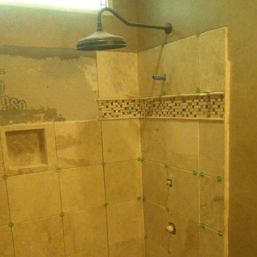 A bathroom shower area under renovation with beige tiles, a tiled accent strip, and a showerhead mounted on the ceiling. Green tile spacers are visible, and some tiles are still being installed.