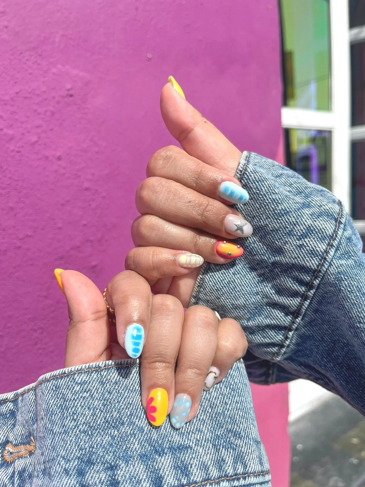 Close-up of two hands with colorful, artistic manicure nails, wearing denim jackets, in front of a pink wall.