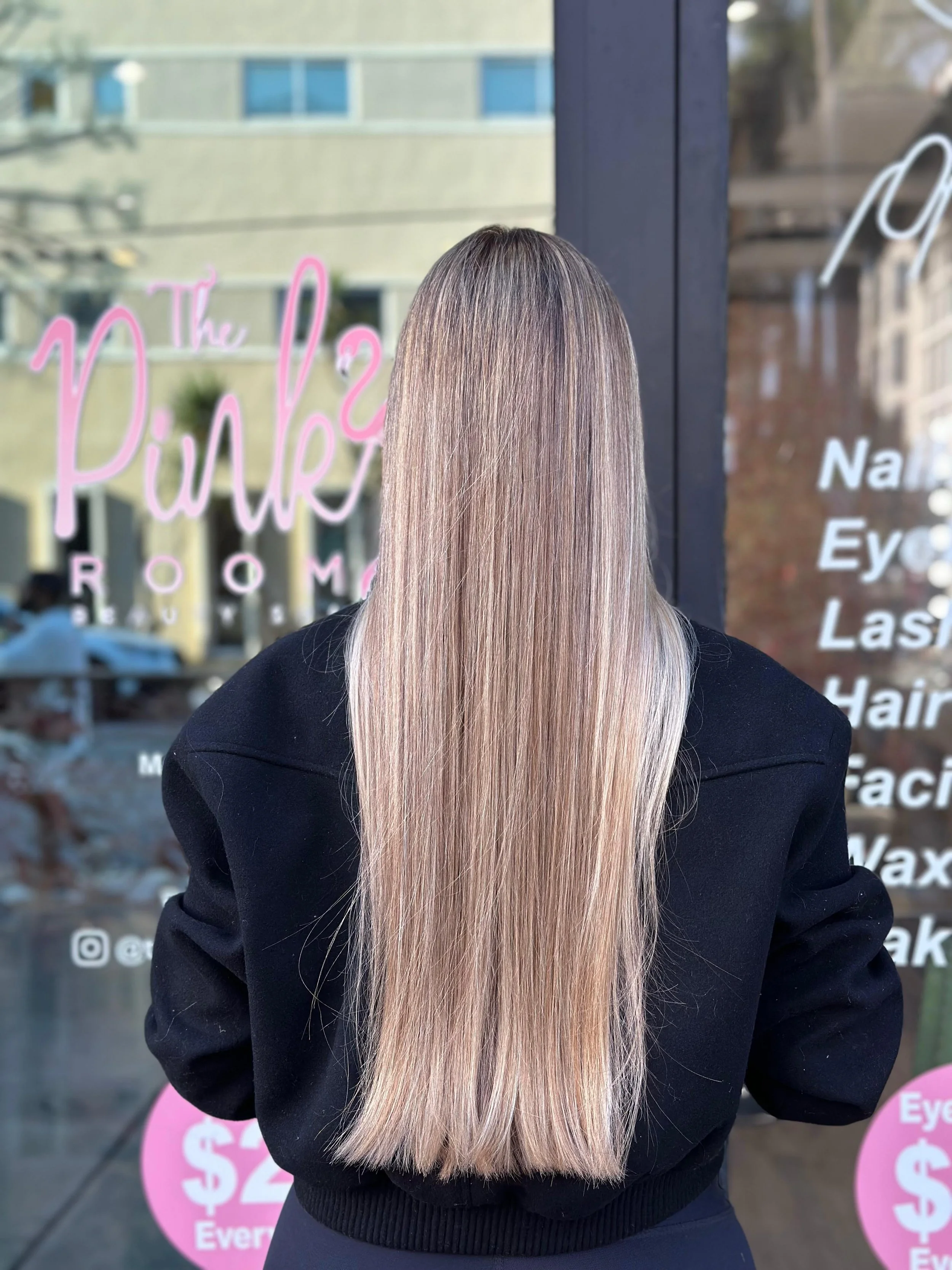 Back view of a woman with long, straight, blonde hair standing outside a salon window.