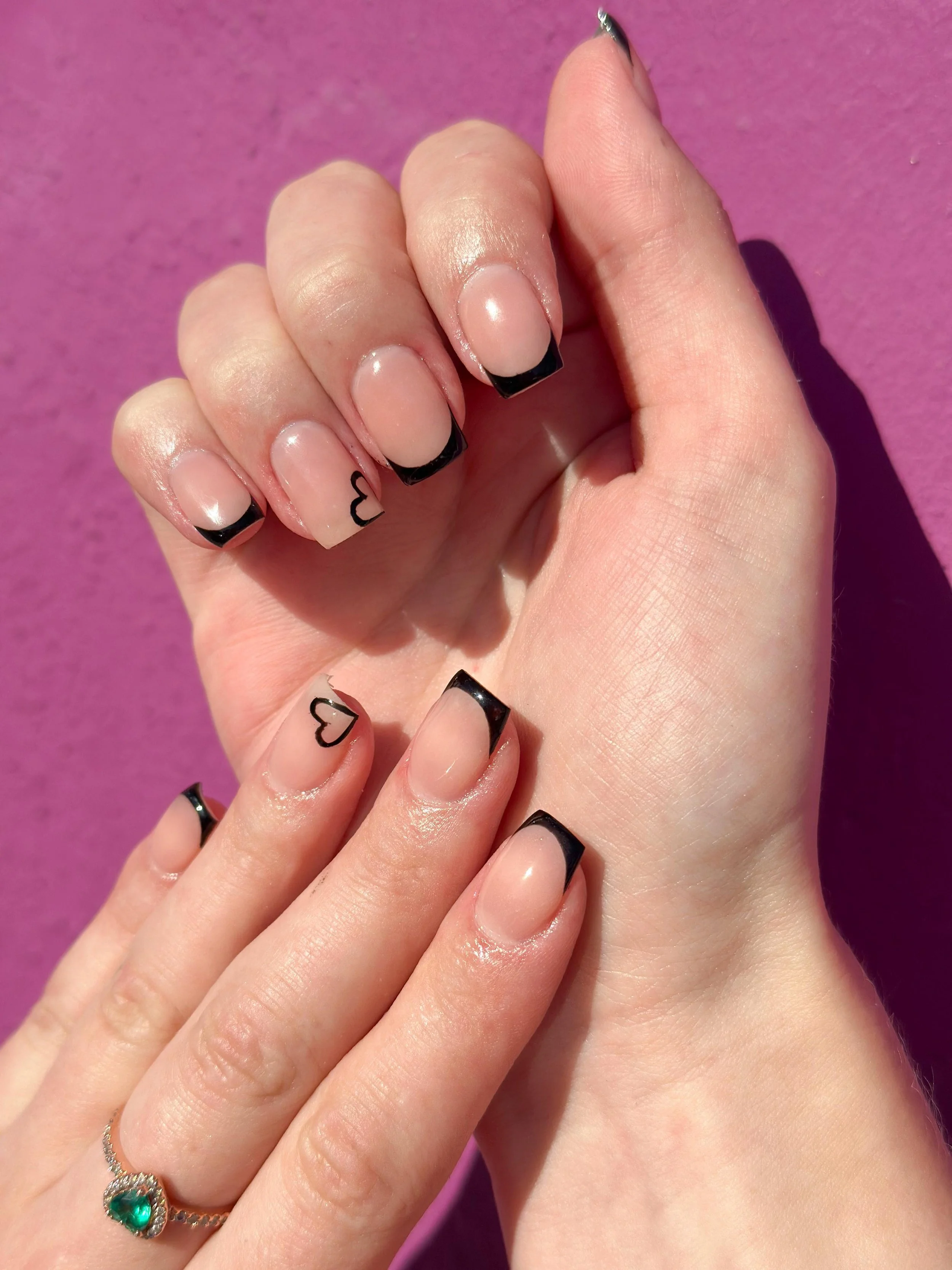 Close-up of two hands with manicured nails featuring black French tips and small heart designs on the ring fingers, held against a pink textured background.
