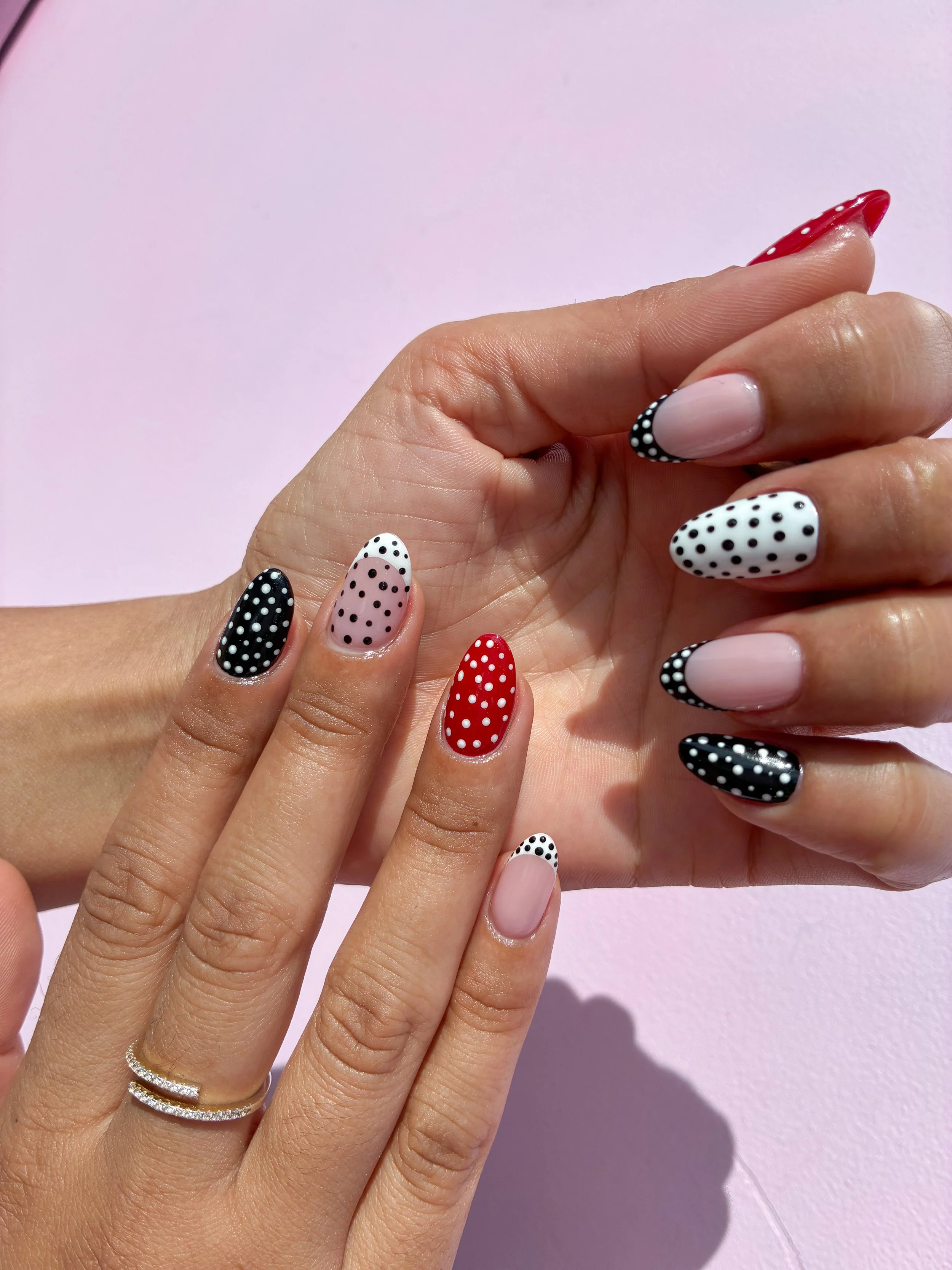 Close-up of two hands with colorful polka dot manicure nails against a pink background.