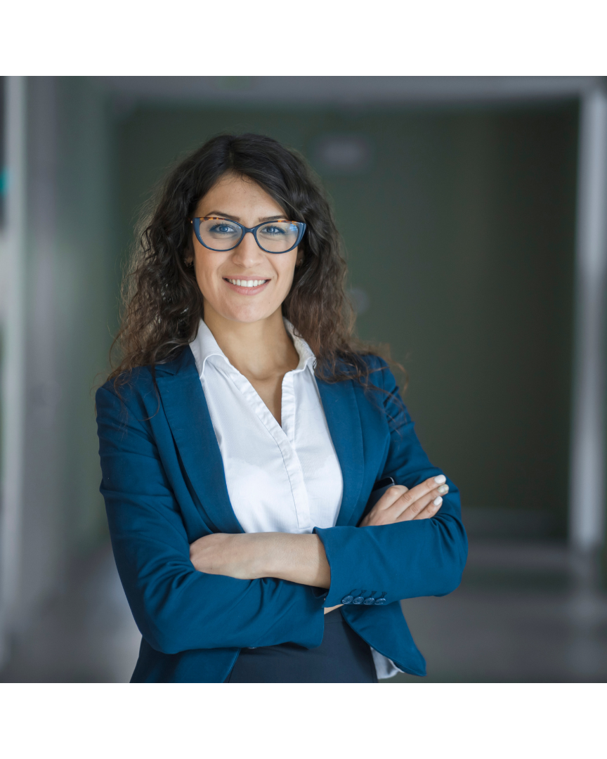 A woman with glasses, long curly brown hair, wearing a white shirt and blue blazer, smiling with arms crossed in a professional setting.