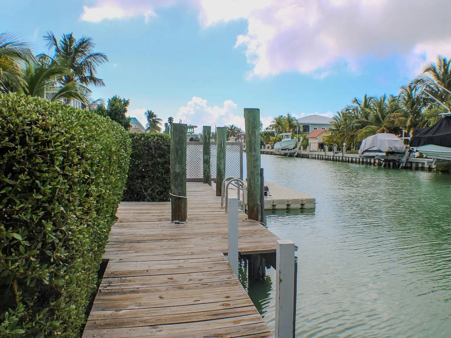 A wooden dock extending over calm water, lined with green bushes on the left and boats docked on the right, with a partly cloudy blue sky overhead.