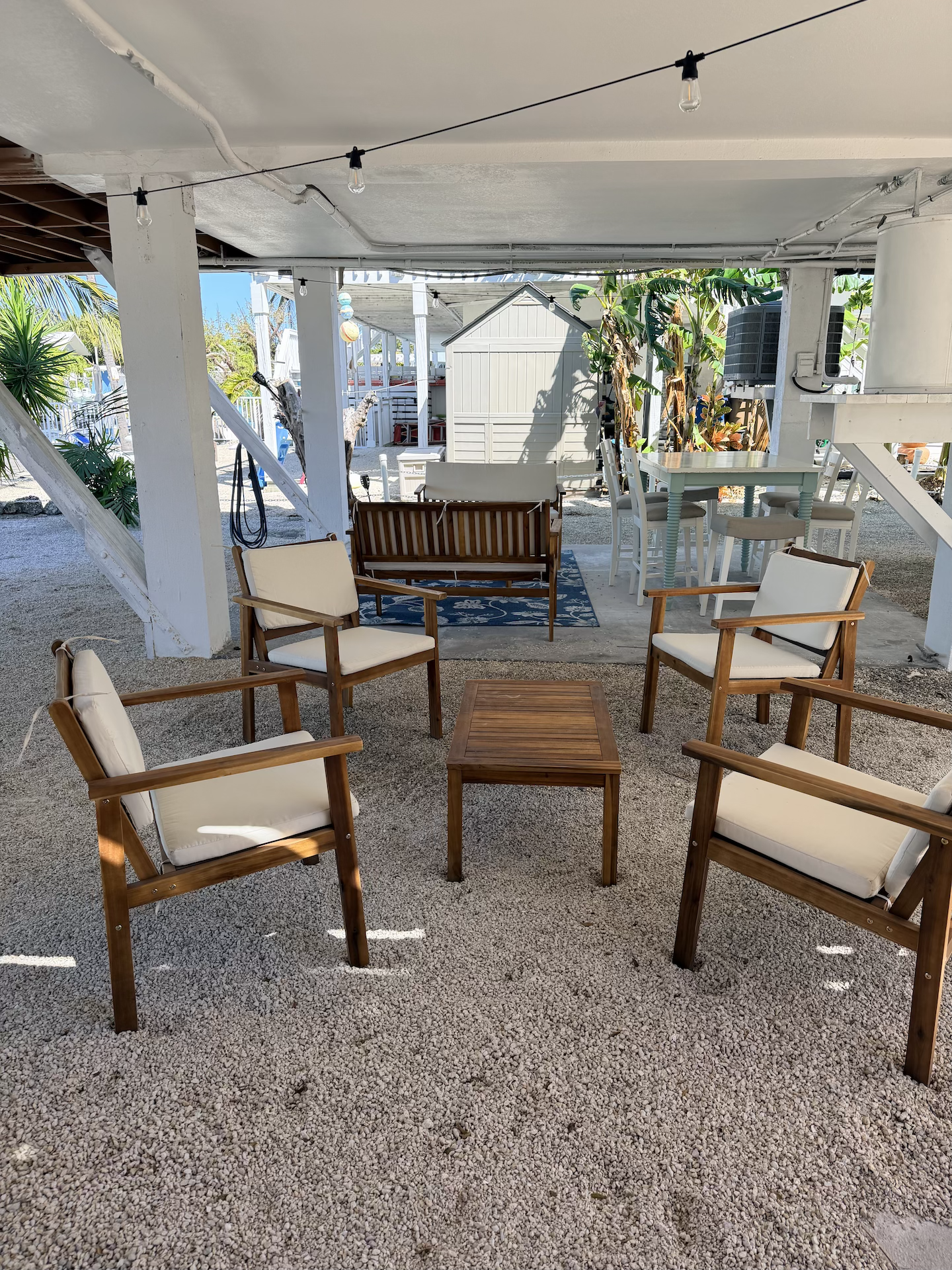 Outdoor patio space with wooden chairs and a small table on gravel, surrounded by plants and a hammock in the background.
