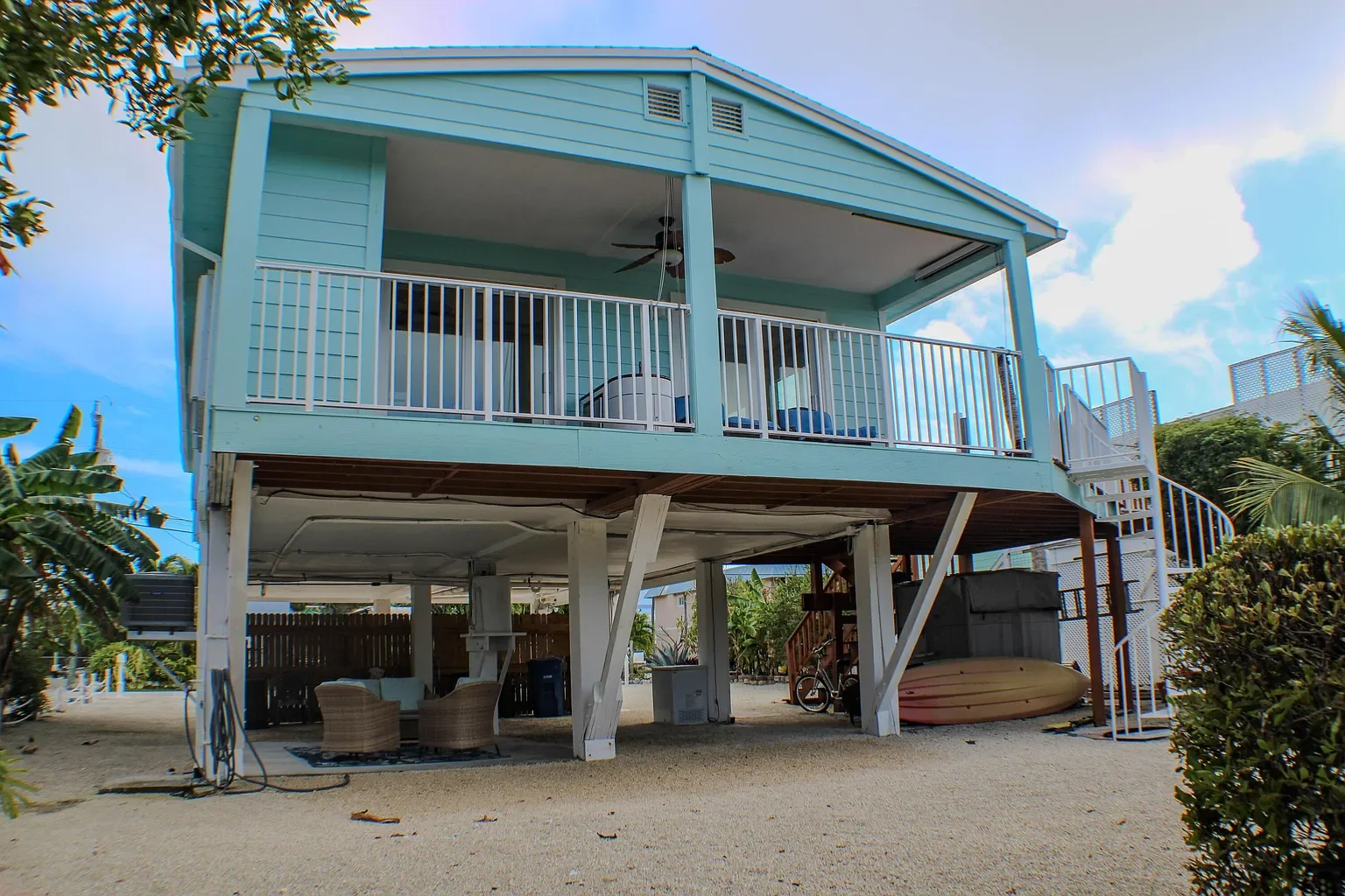 A two-story beach house painted in light blue with a wraparound porch, a ceiling fan, and outdoor furniture underneath. There is a staircase on the right side of the house, a kayak stored underneath, and tropical plants surrounding the house.